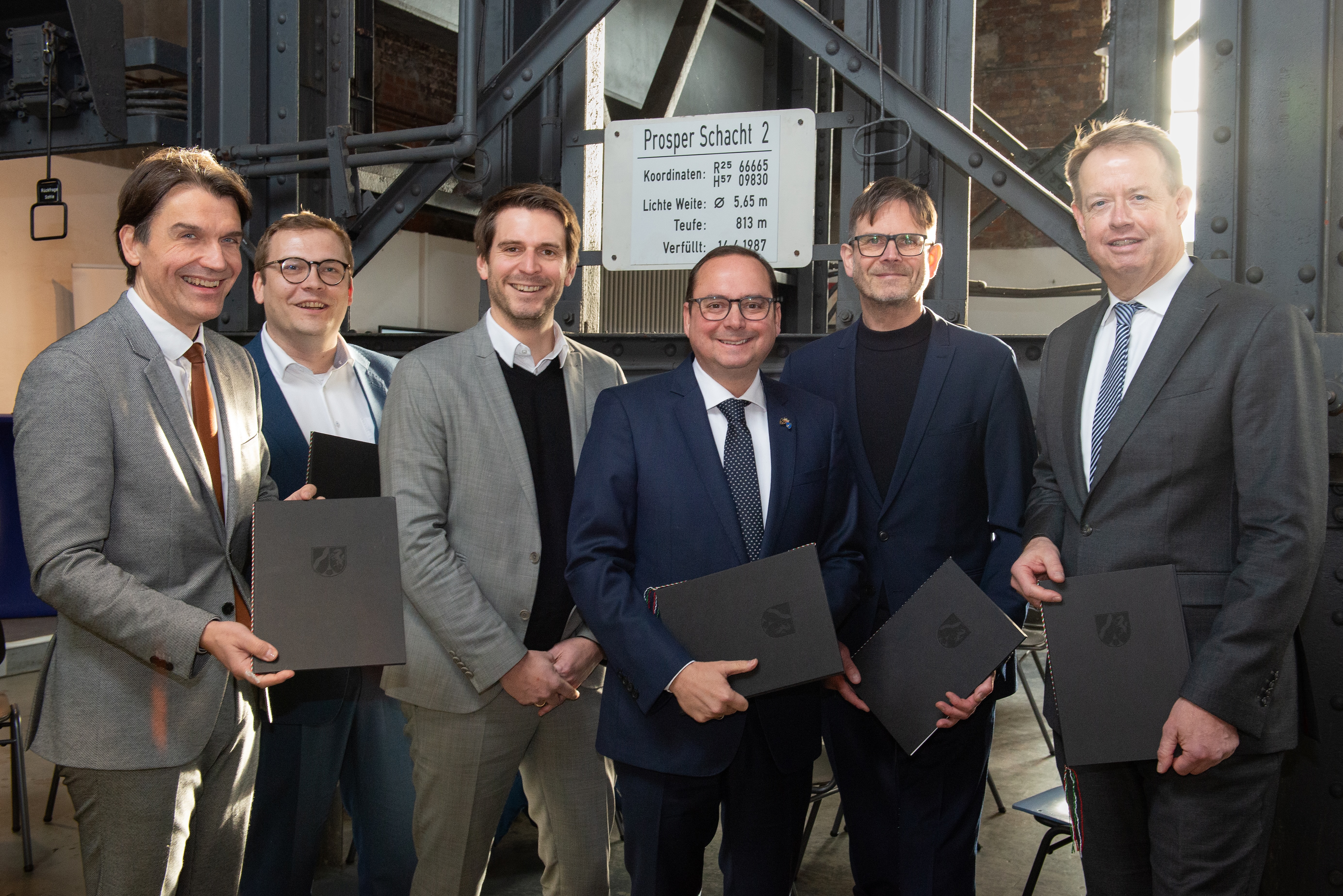 6 Männer lächeln für ein Gruppenfoto in die Kamera und haben die Bescheidübergaben in den Händen.