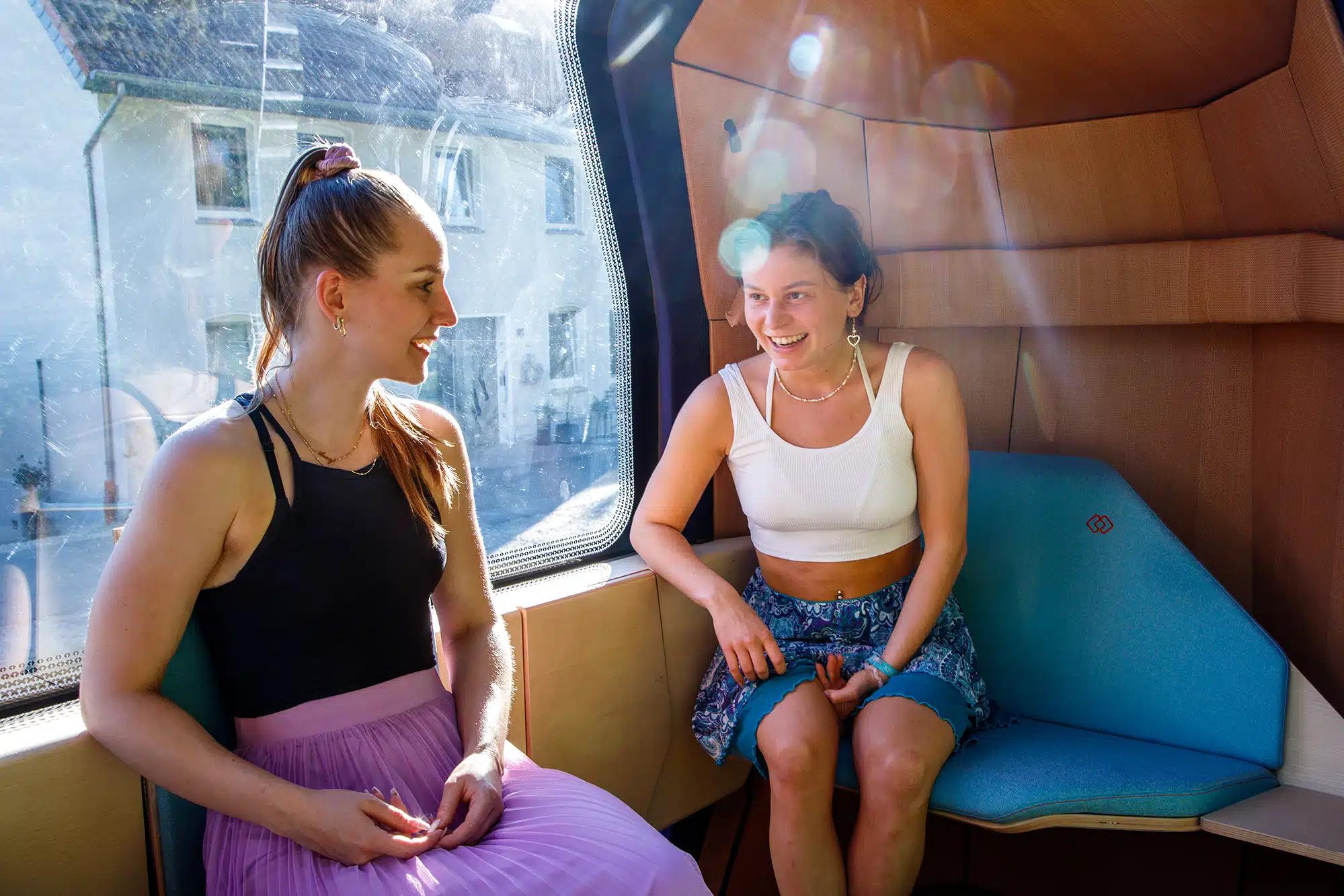 Zwei Frauen sitzen in selbstfahrender Kabine auf Schienen, die einem kleinen Zug-Wagon ähnelt