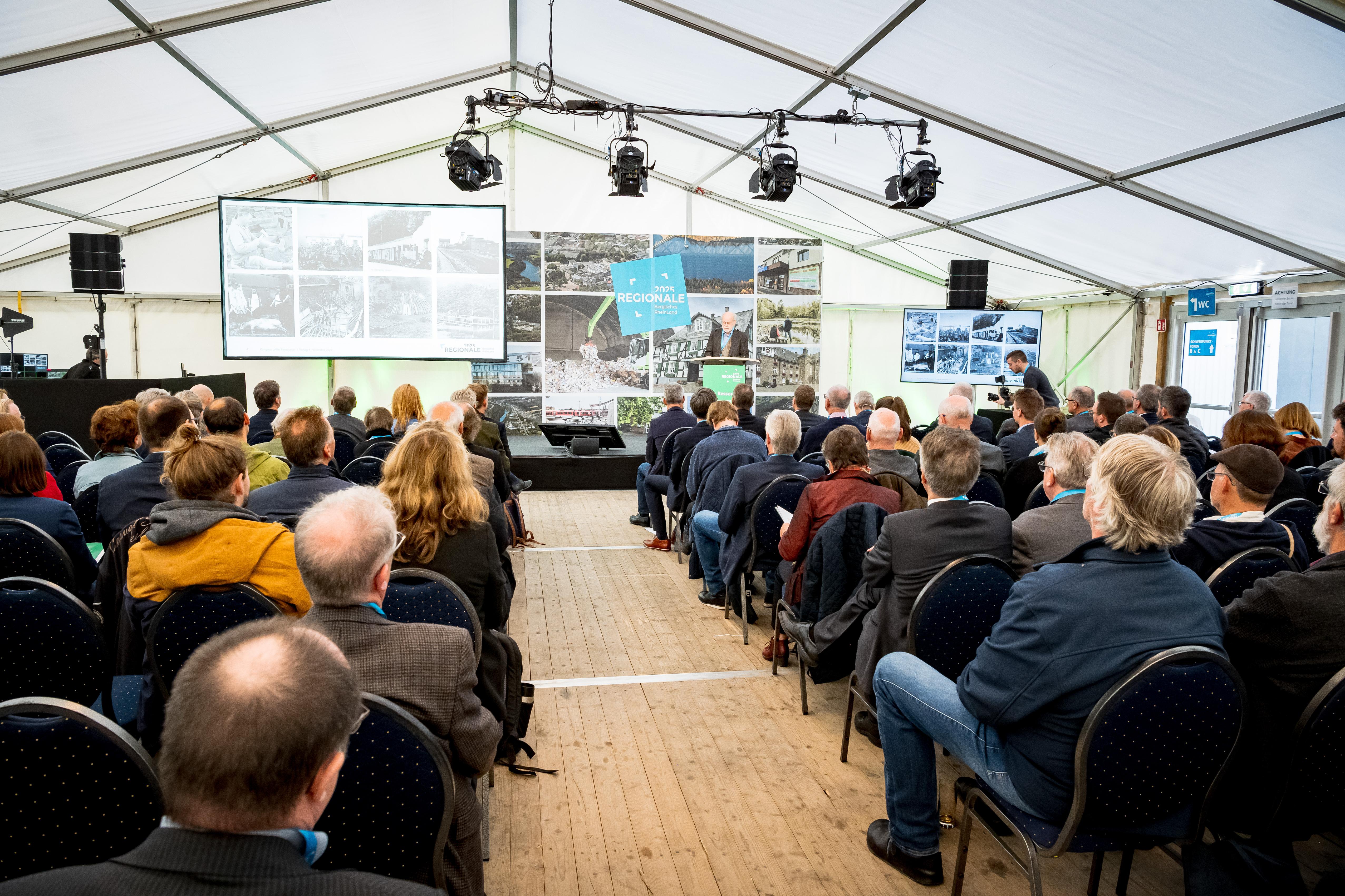 Es ist ein Konferenzraum mit Stuhlreihen abgebildet, auf denen viele Menschen sitzen.