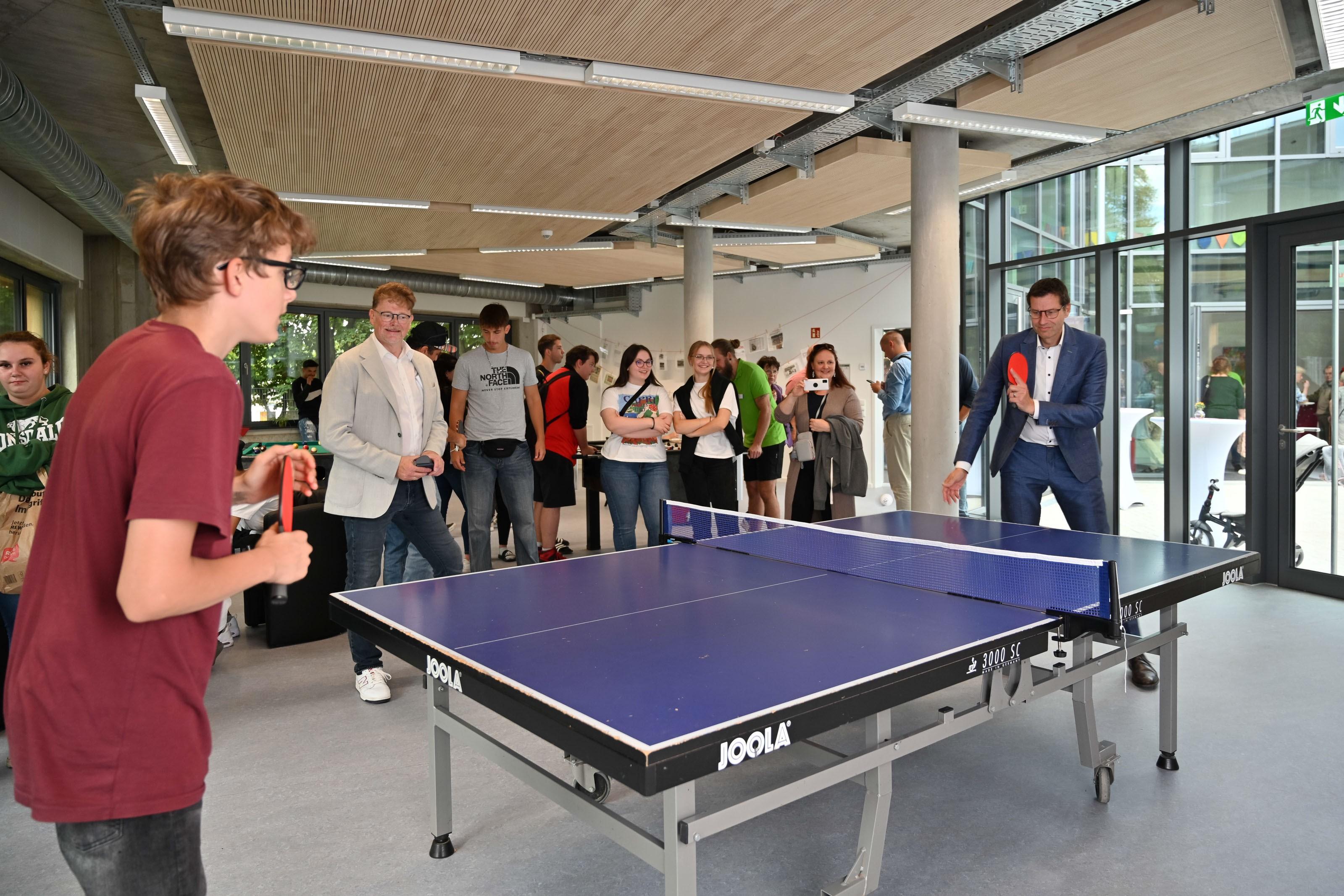 Es spielen zwei Personen gegeneinander Tischtennis. Um die Platte stehen einige Personen, die zuschauen.