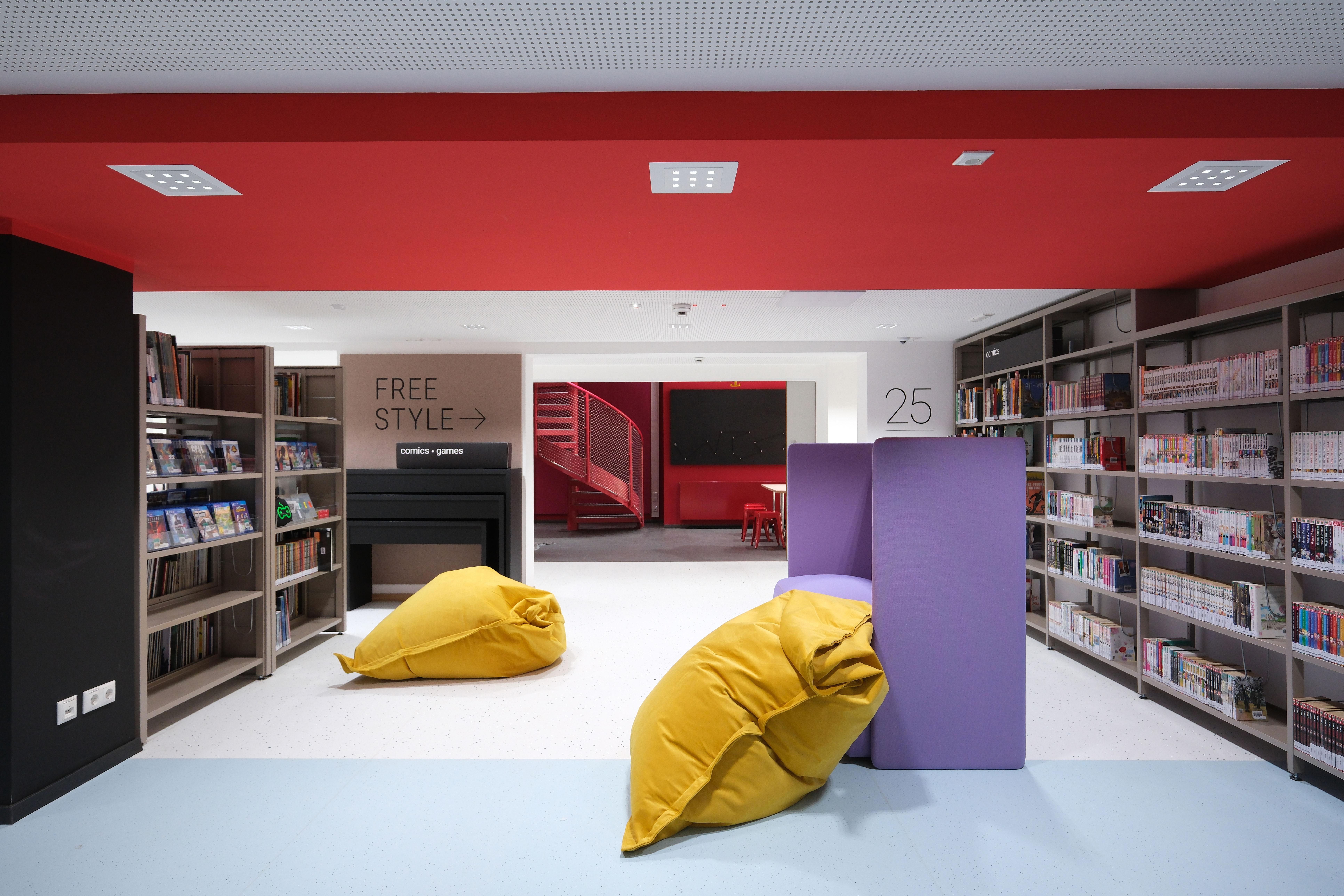 Es sind Regale mit vielen Büchern zu sehen. Zudem liegen Sitzkissen im Raum.