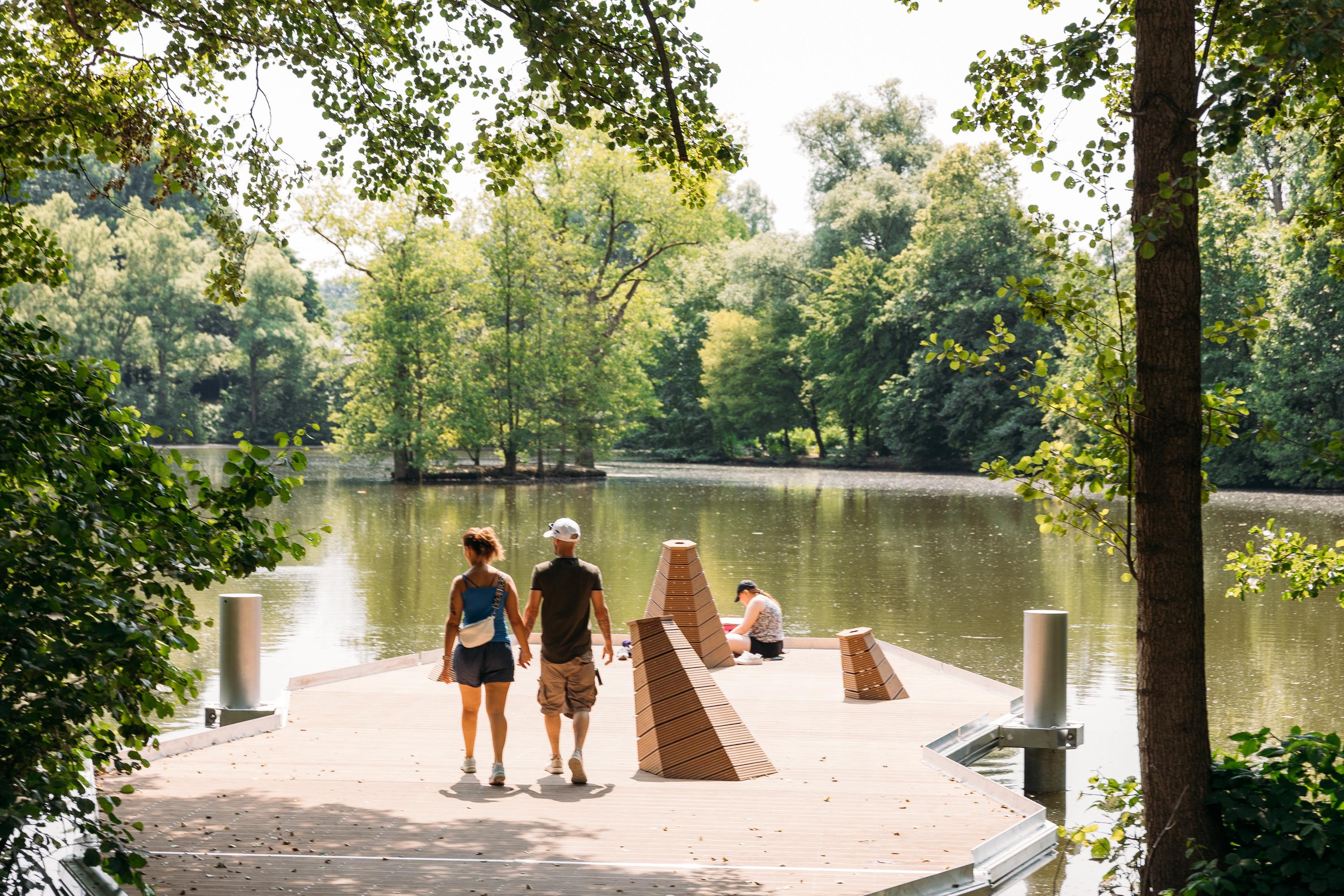 Auf einer kleinen, schwimmenden Plattform in einem See, um dessen Ufer herum Bäume wachsen, sitzt eine Person direkt am Wasser. Zwei weitere Personen gehen Hand in Hand über die Plattform und schauen auf den See.