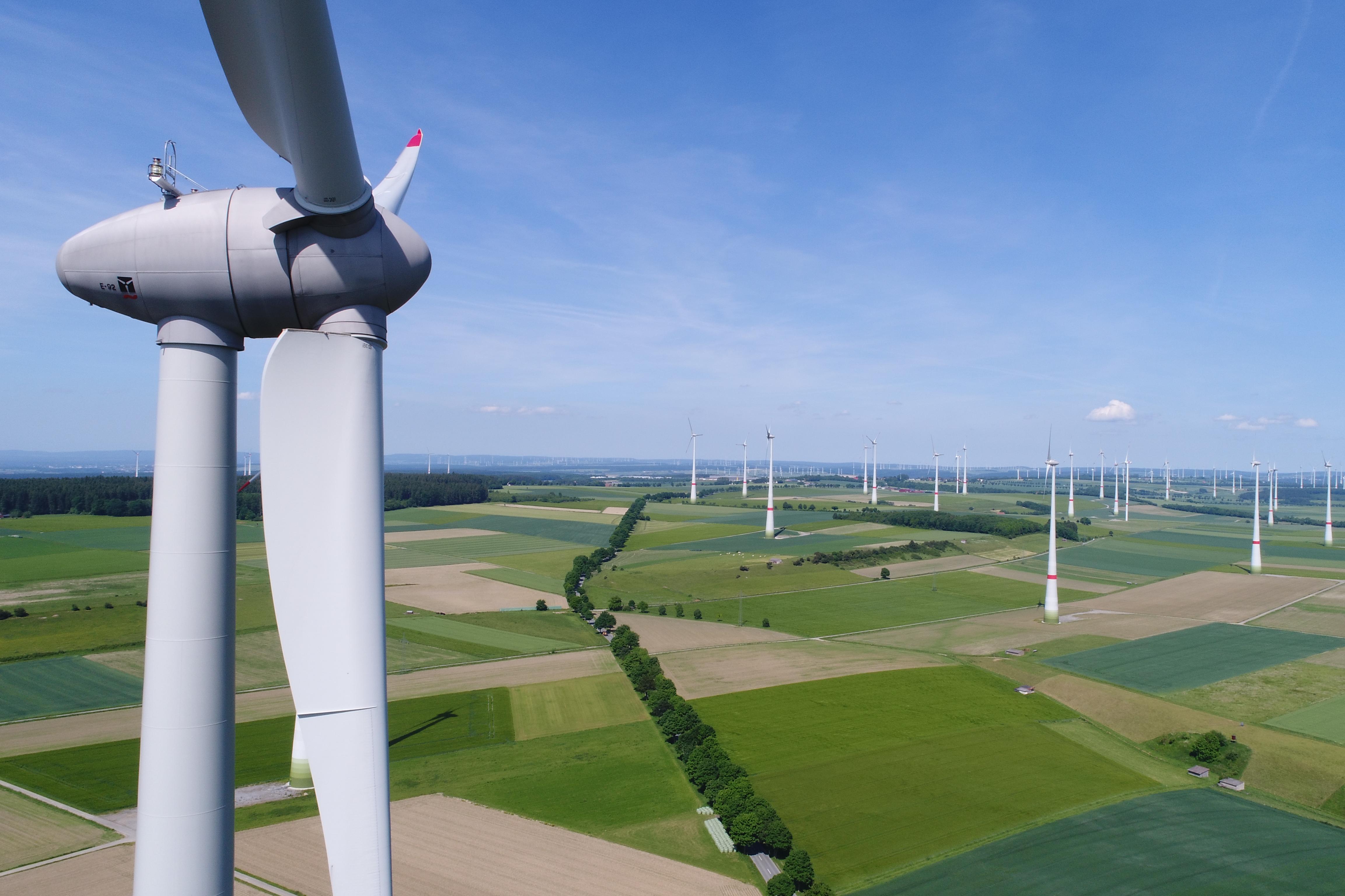 Ein Windrad steht in einer freien Fläche grüner Felder. Der Himmel ist blau, am Horizont sind weitere Windräder zu erkennen.
