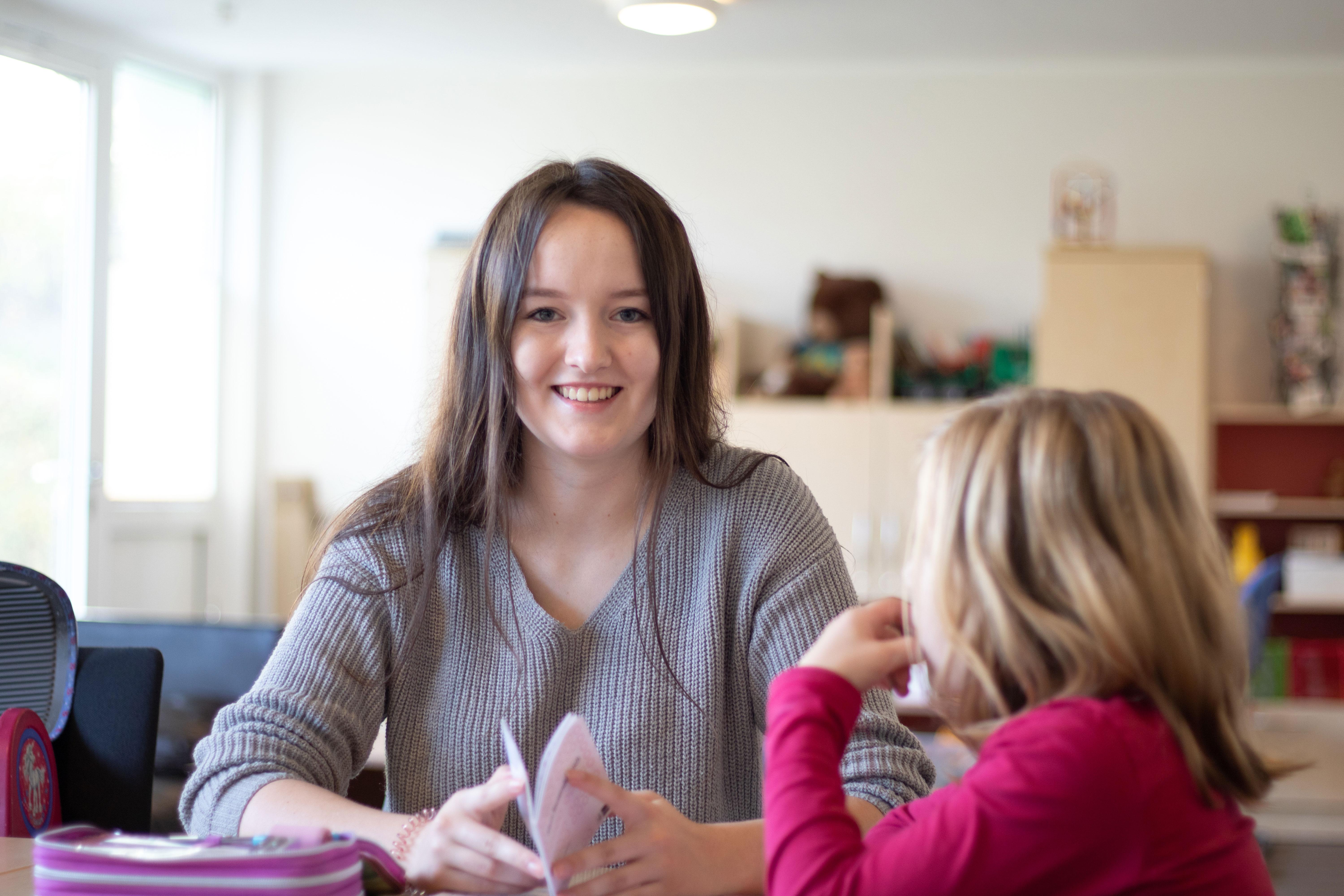 Eine junge Frau mit langen braunen Haaren sitzt an einem Tisch und lächelt. Sie zeigt einem Kind, das ihr gegenüber sitzt, ein Heft.