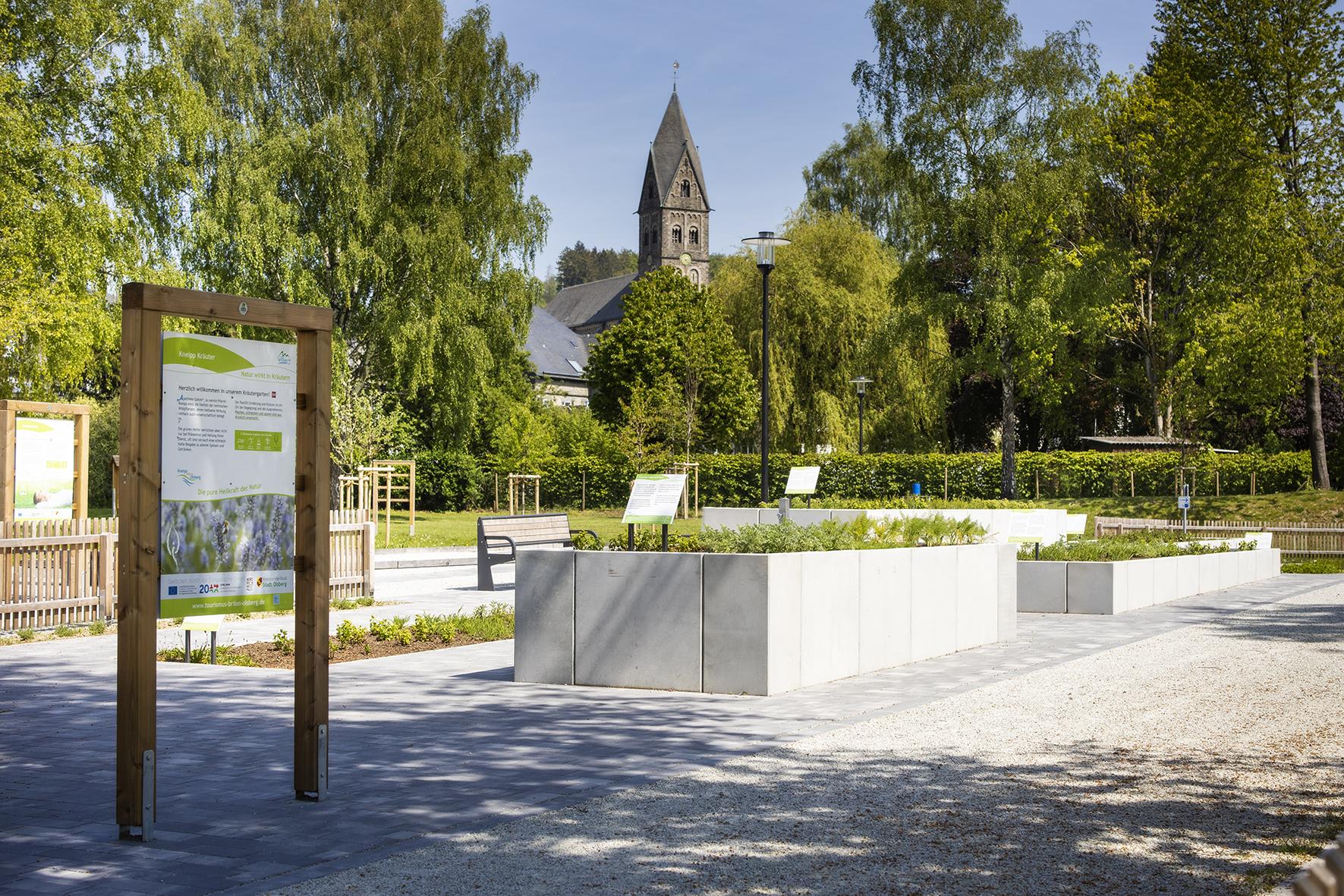 Ein begrünter Dorfplatz, von Bäumen umringt und mit Beeten in verschiedenen Höhen. Im Hintergrund ragt ein Kirchturm hervor. Vor dem Platz steht ein Infoschild.
