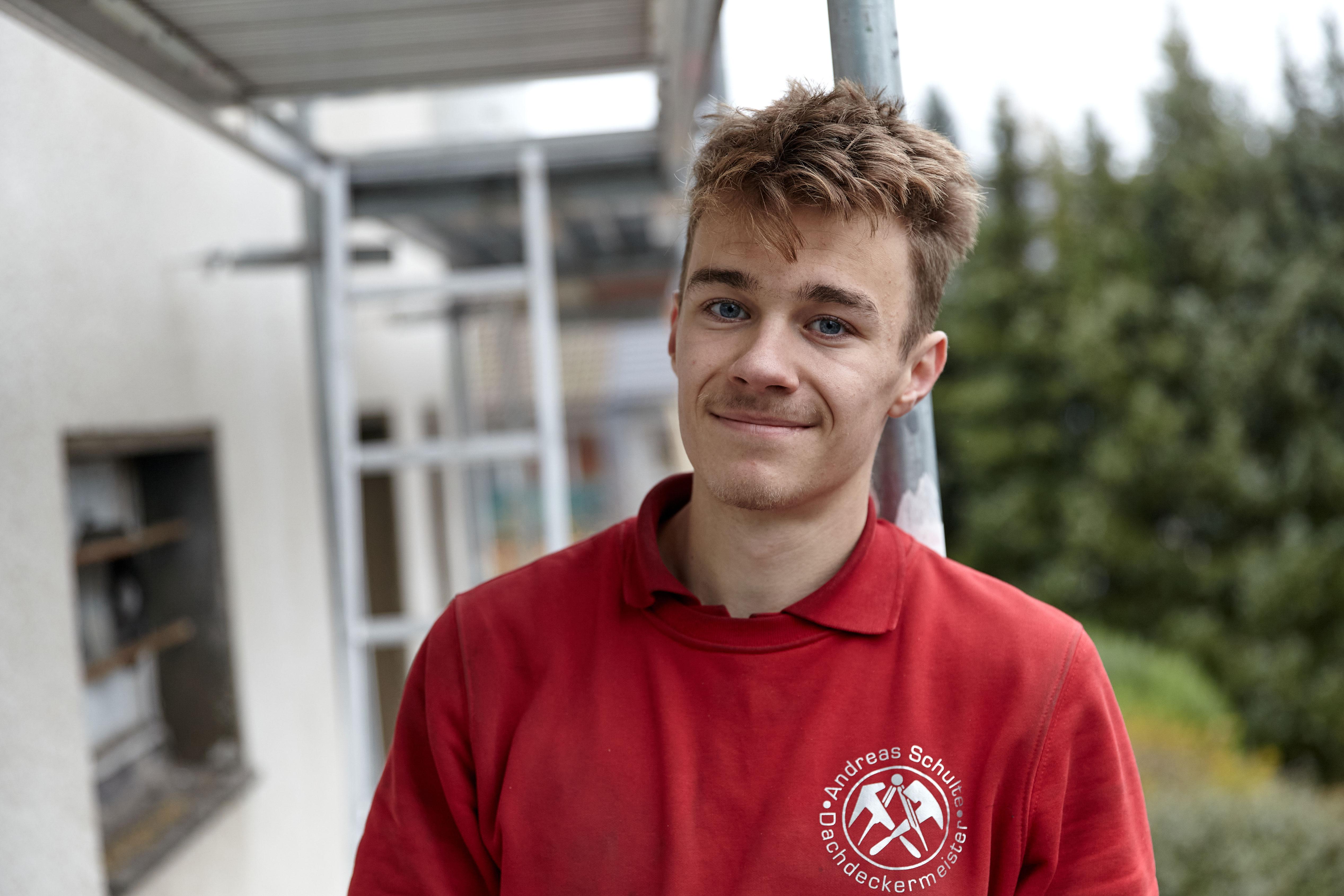 Ein junger Mann in rotem Poloshirt mit dem Logo einer Dachdeckerfirma lehnt an einem Gerüst und lächelt in die Kamera. 