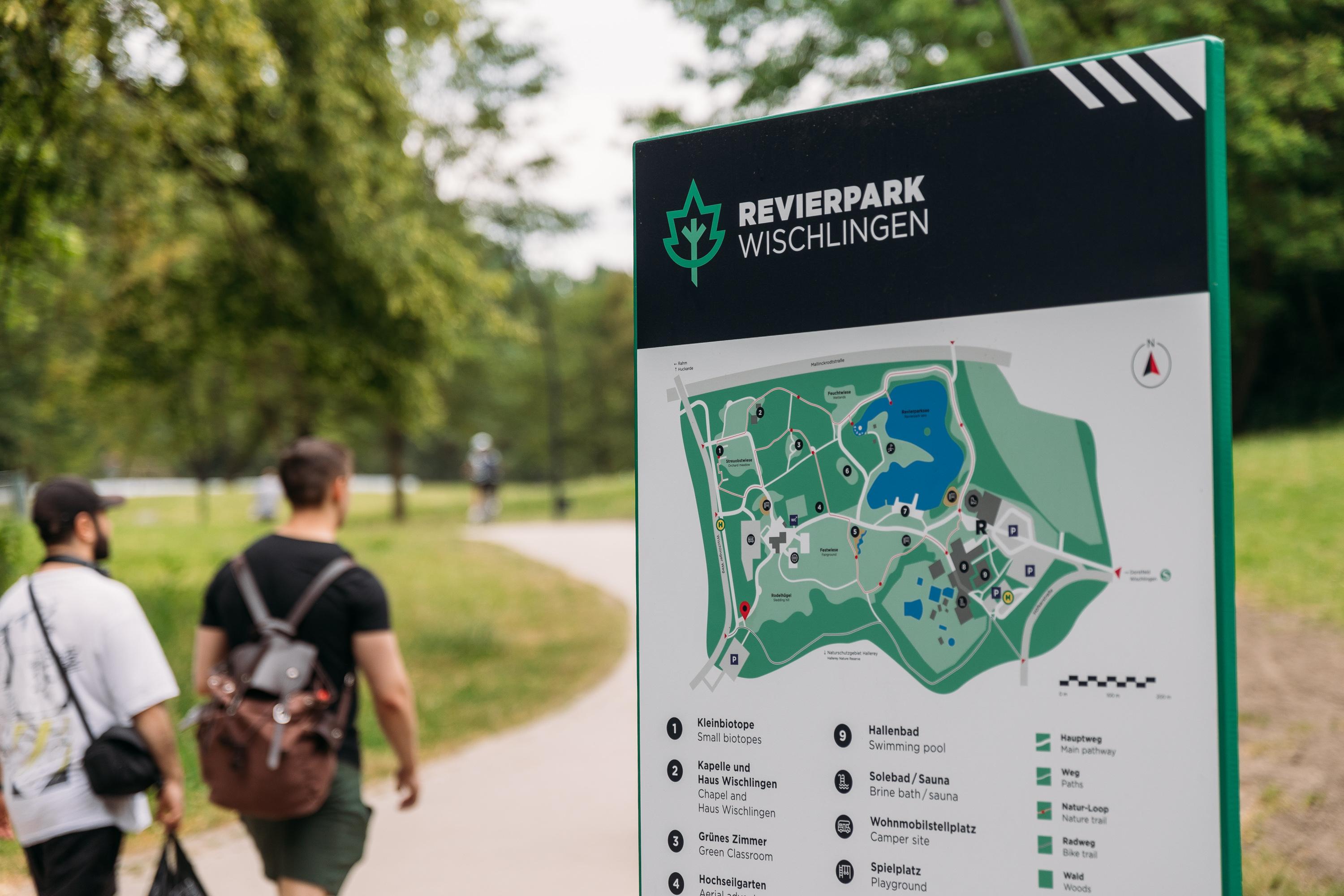 Zwei sportlich gekleidete Männer gehen auf einem Wanderpfad, an dessen Seite ein Schild steht, auf dem die verschiedenen Routen abgebildet sind.