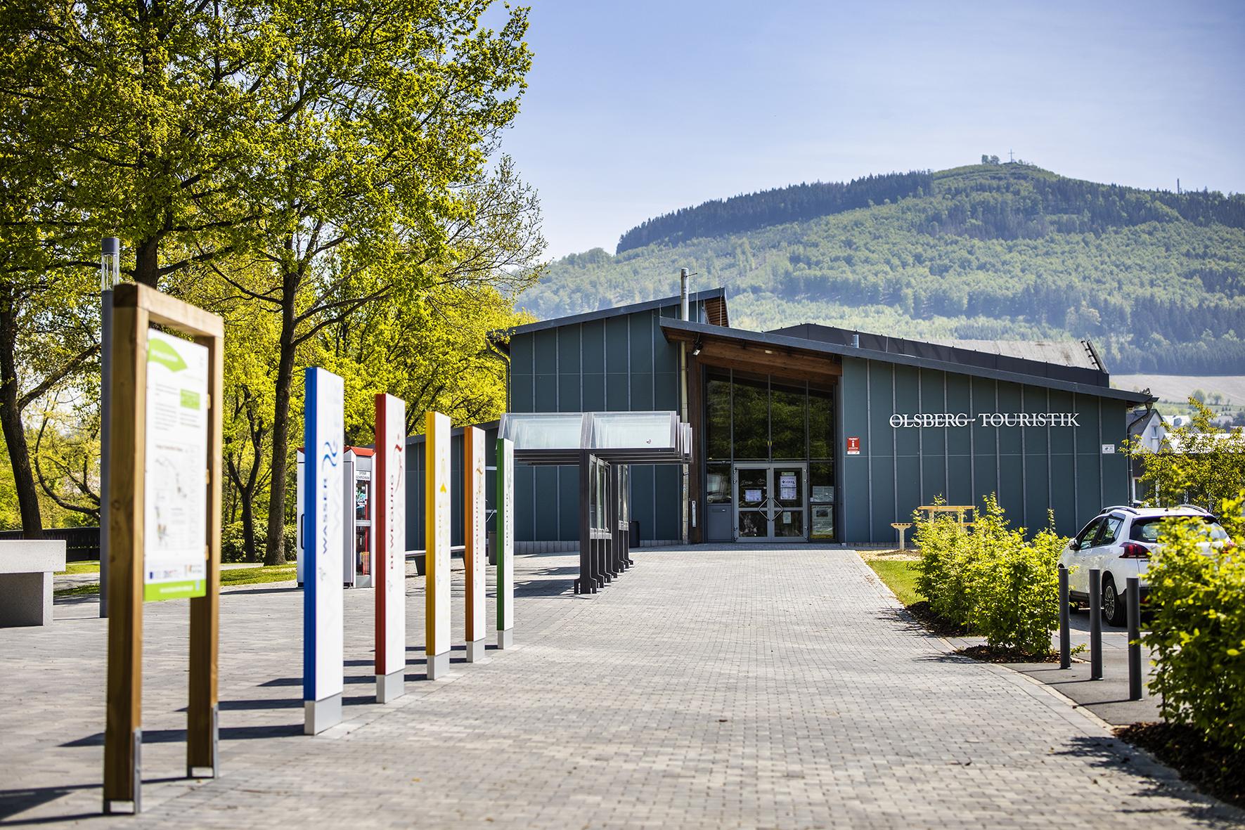Die Tourismus-Information Olsberg. Auf dem Platz vor dem Gebäude stehen Infotafeln, im Hintergrund bewaldete Hügel.