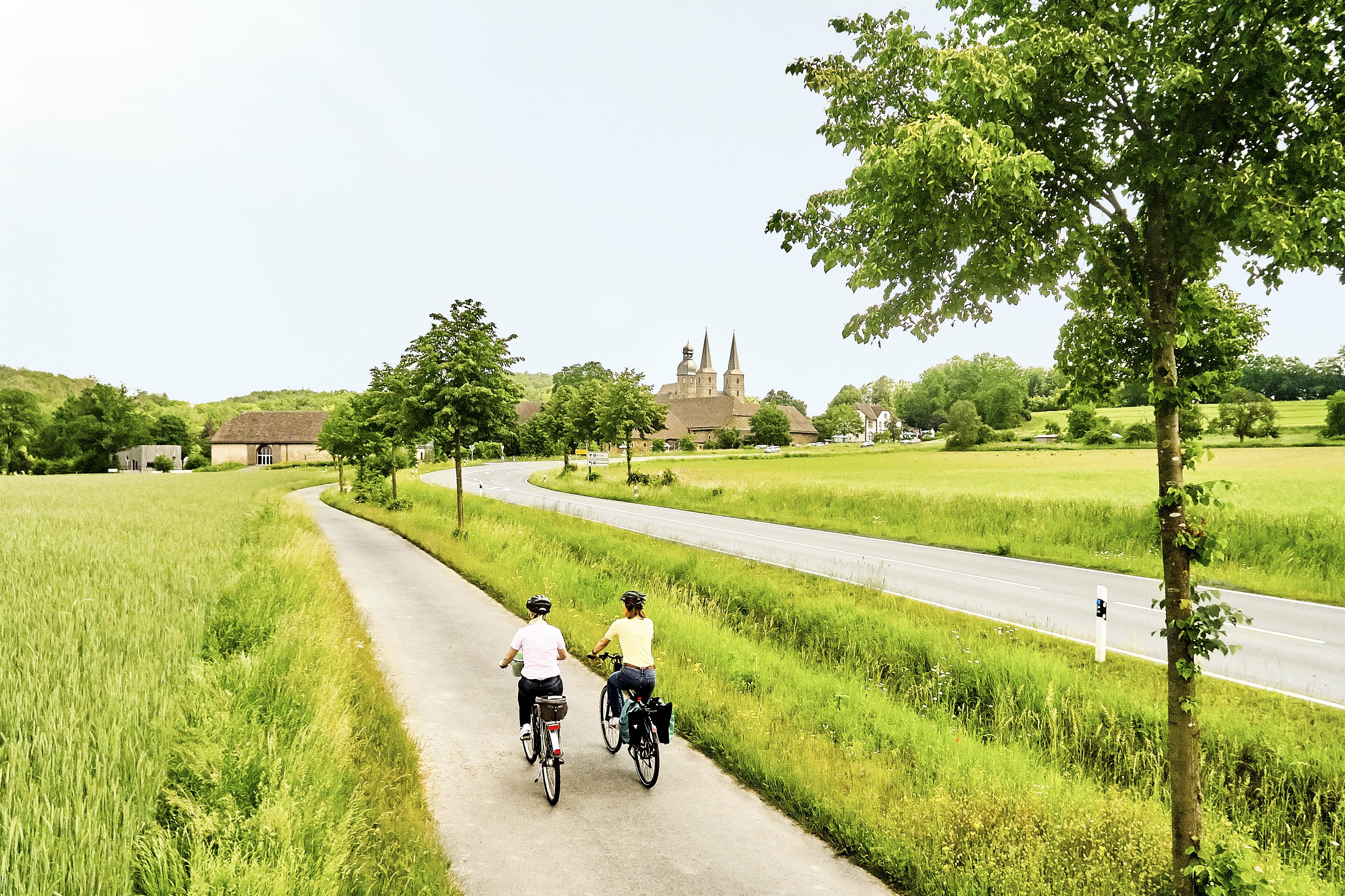 Ein Fahrradweg führt an einer Landstraße entlang über grüne Felder. Auf dem Weg radeln zwei Menschen, am Horizont ein Dorf. 