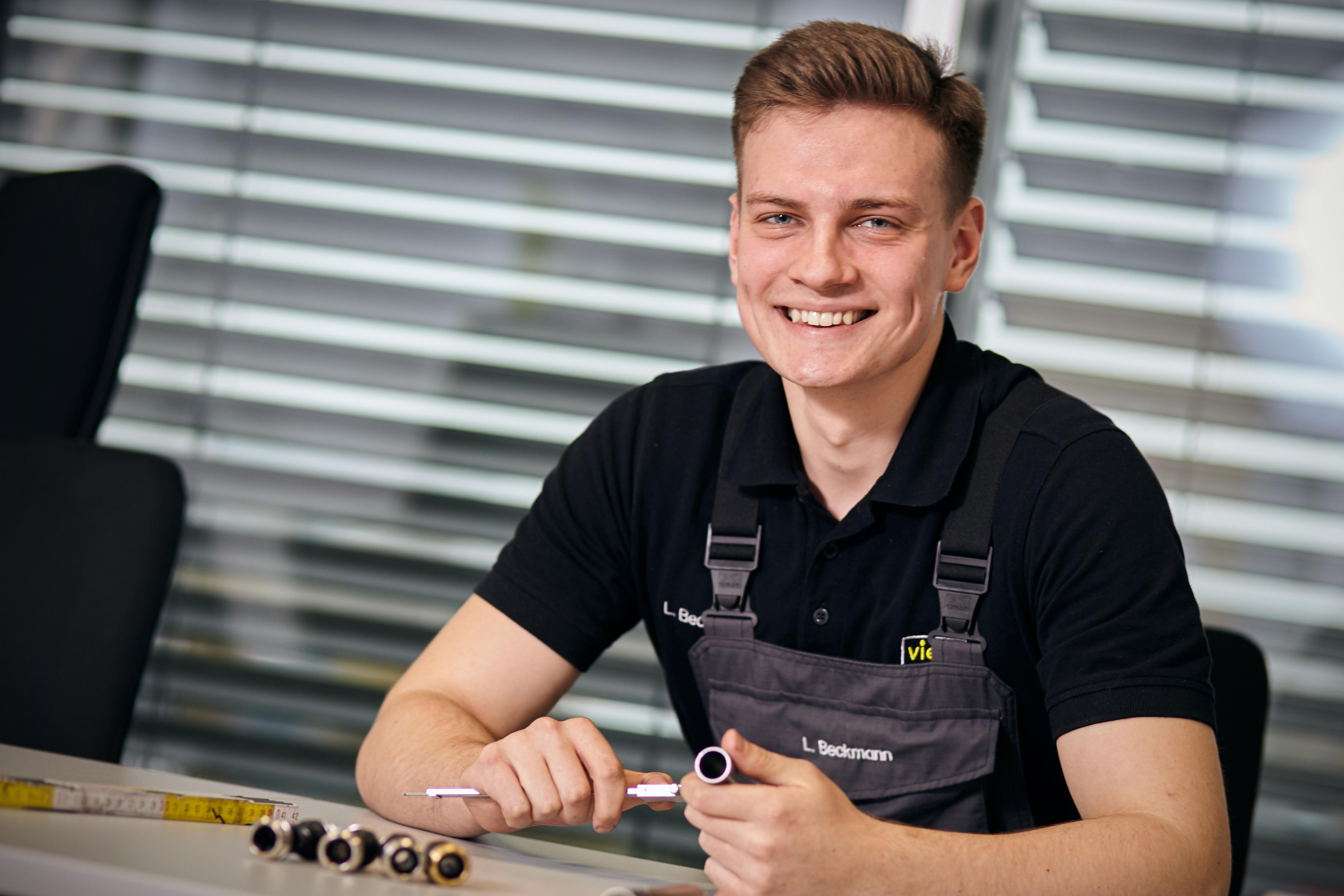 Ein junger Mann in Arbeitskleidung sitzt an einem Tisch, auf dem verschiedene Bauteile liegen. Er lächelt in die Kamera und schraubt dabei an einem Bauteil.