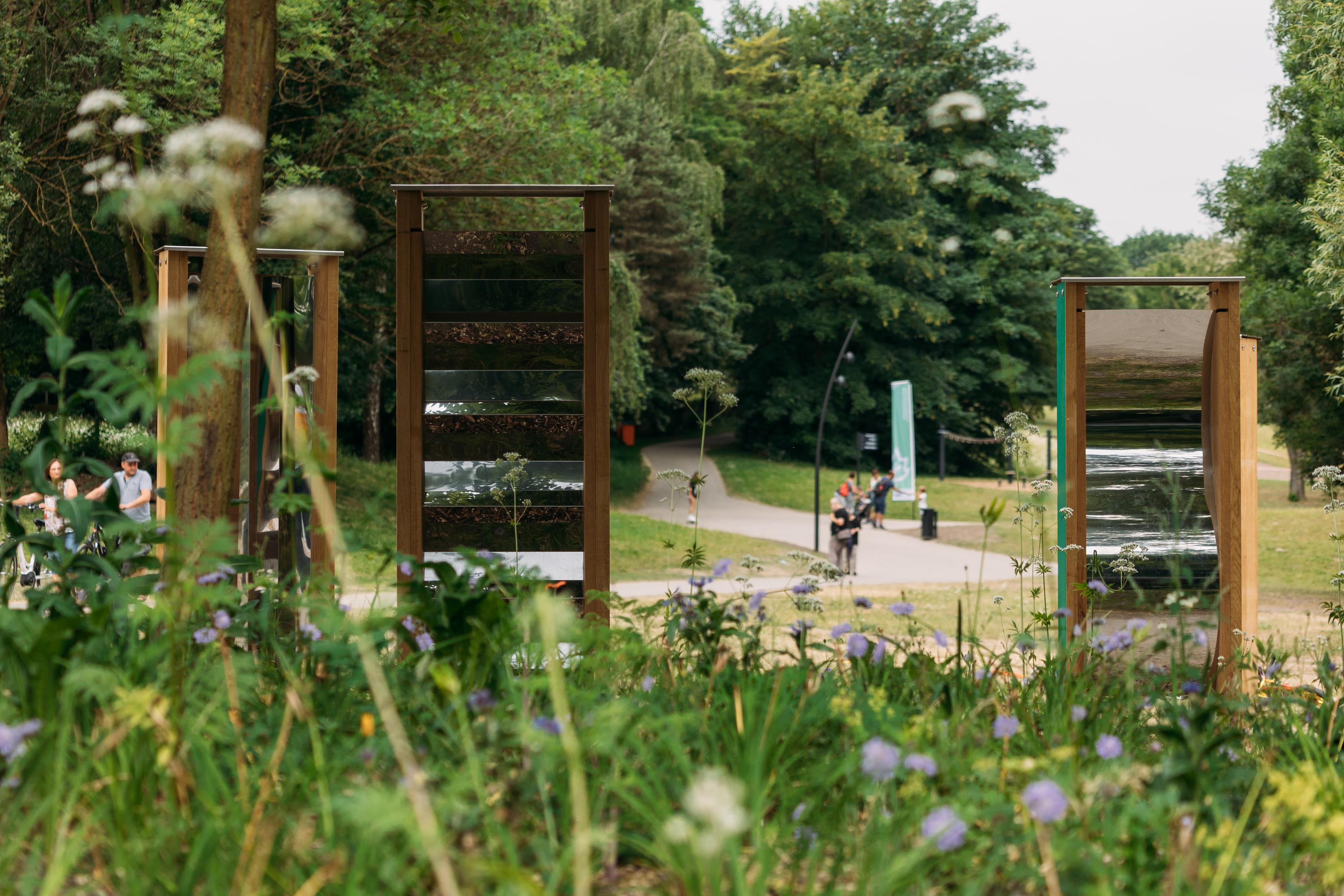 An einem Punkt, an dem mehere Wege zusammenführen wachsen hochrankende Wildblumen. Im Hintergrund sind Spaziergänger und Fahrradfahrer zu sehen.