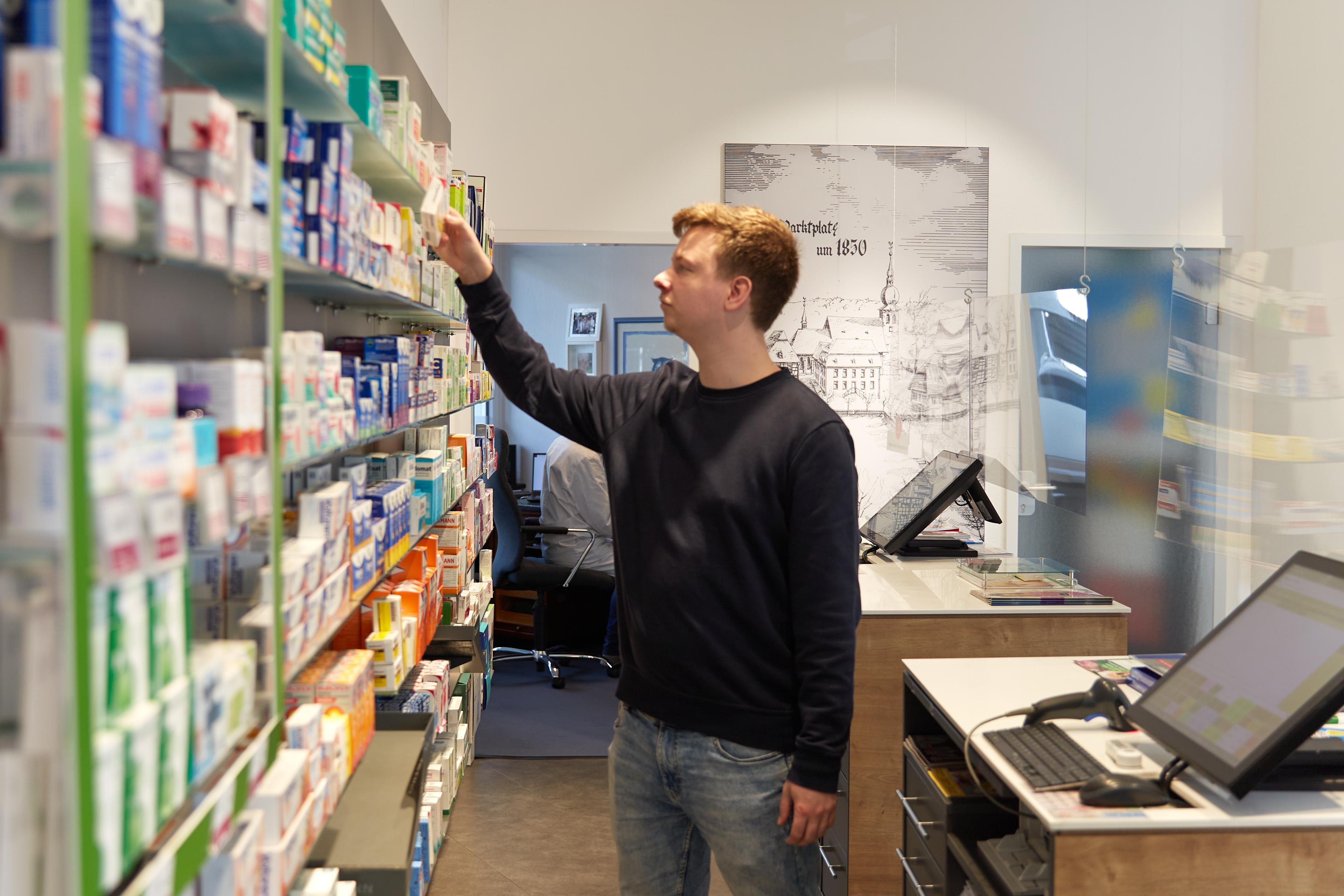 Ein junger Mann steht in einer Apotheke an einem Regal hinter der Kasse und sucht nach einem Medikament.