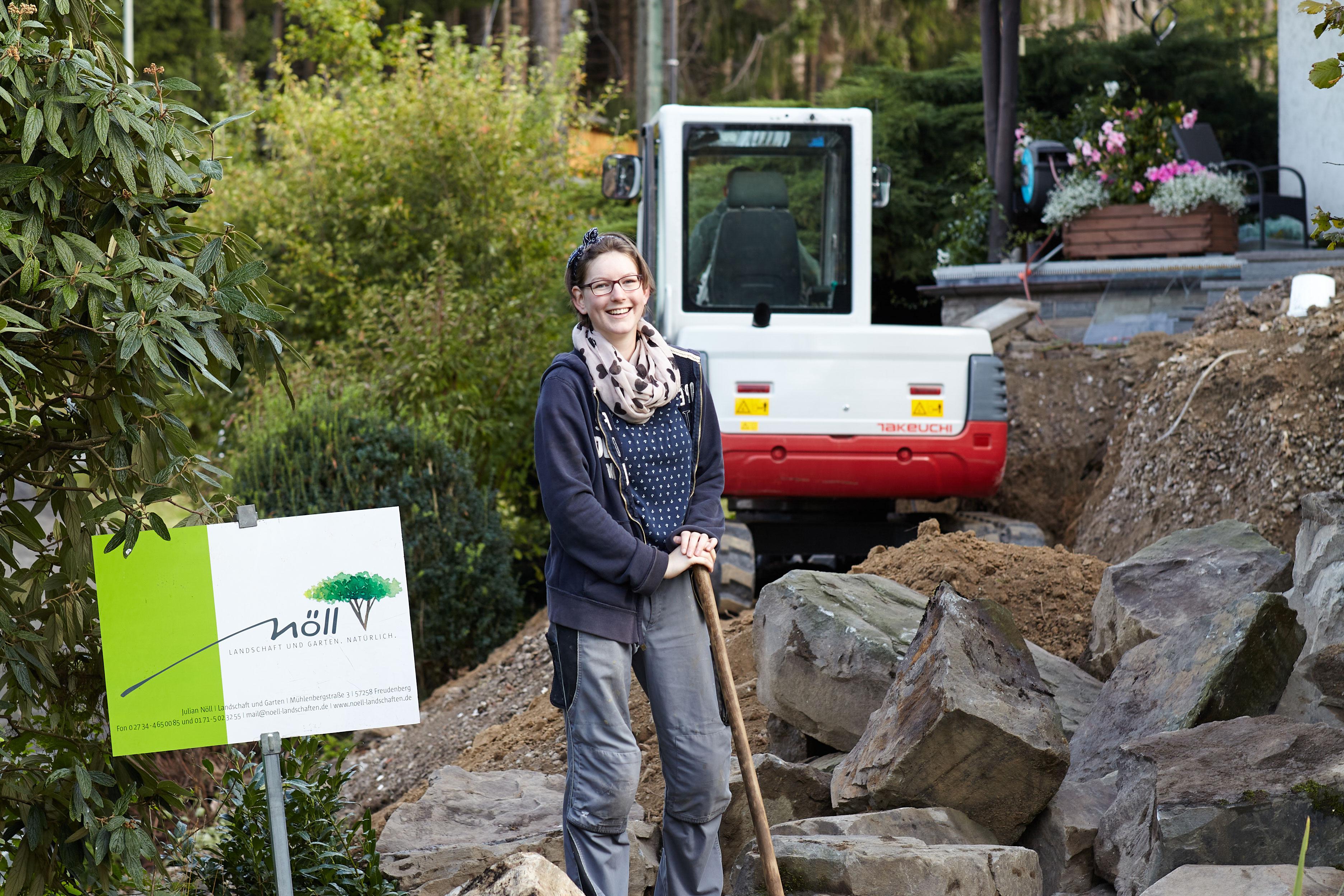 Eine junge Frau steht in einem aufgebrabenen Garten. Sie lächelt in die Kamera und hält eine Schaufel in der Hand.