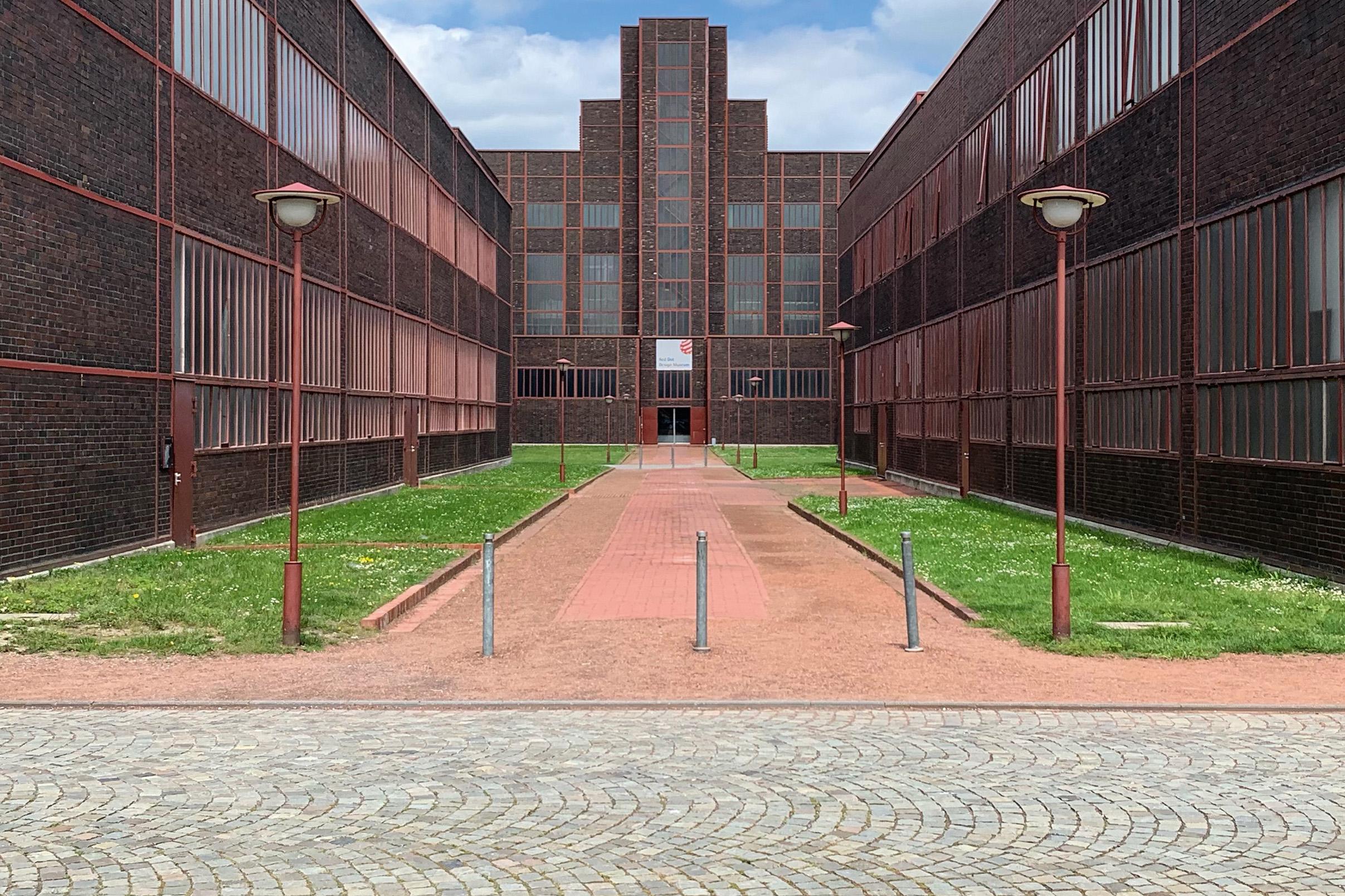 Ein Klinkerbau von außen mit roten Details und Straßenlaternen. Zwischen den einzelnen Gebäudeteilen verlaufen begrünte Fußwege.