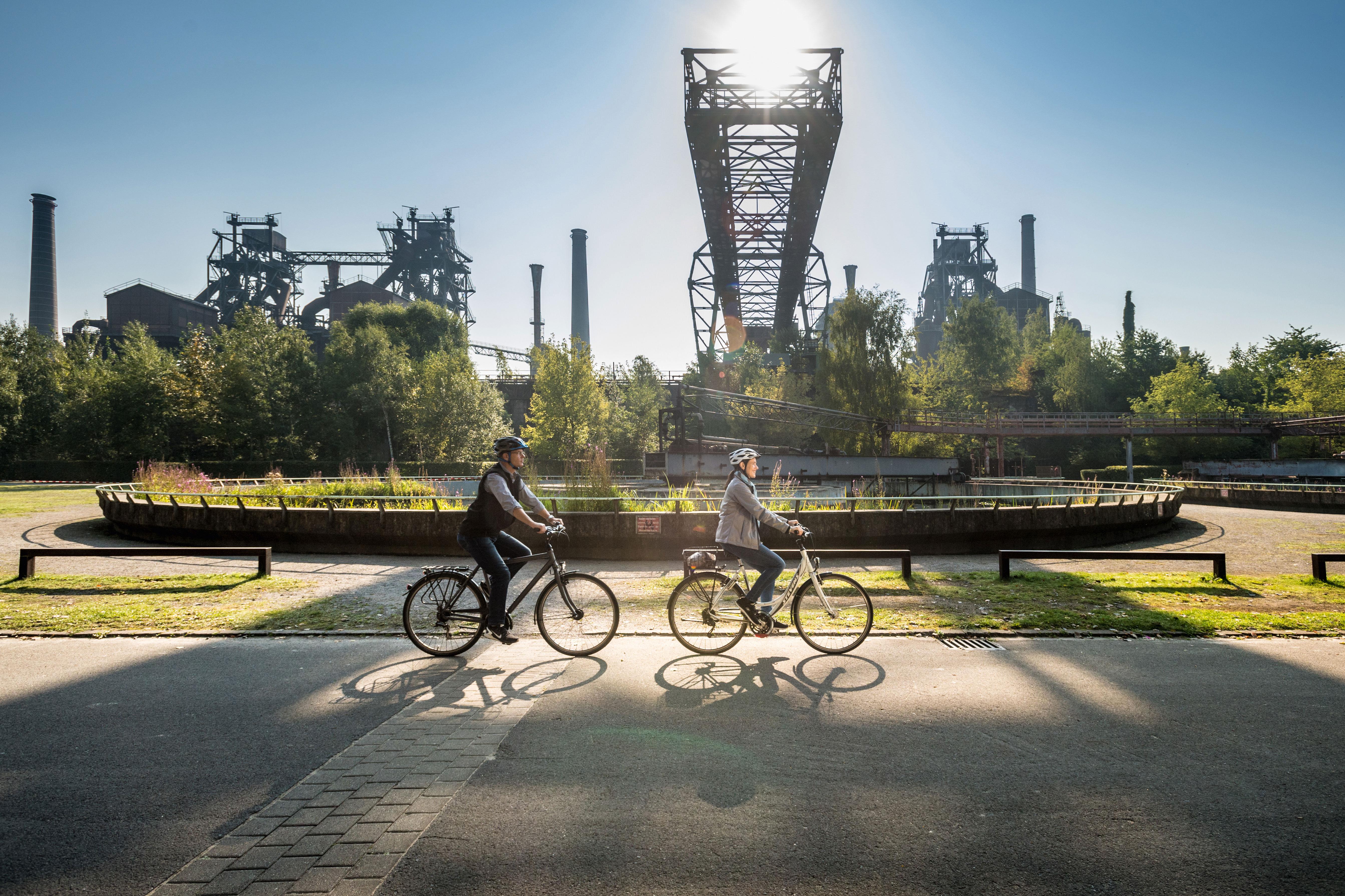 Zwei Personen radeln auf einem Fahrradweg, der an einer alten Industrieanlage vorbeiführt.