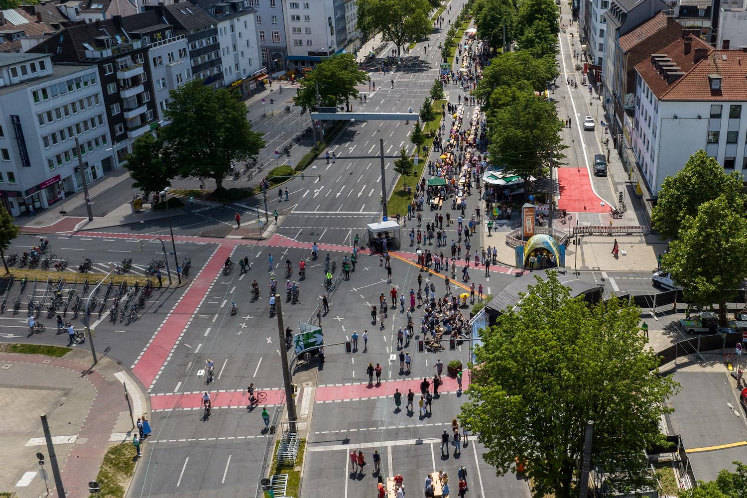 Eine belebte Straßenkreuzung aus der Vogelperspektive. Auf der Straße halten sich viele Menschen auf, während keine Autos unterwegs sind.