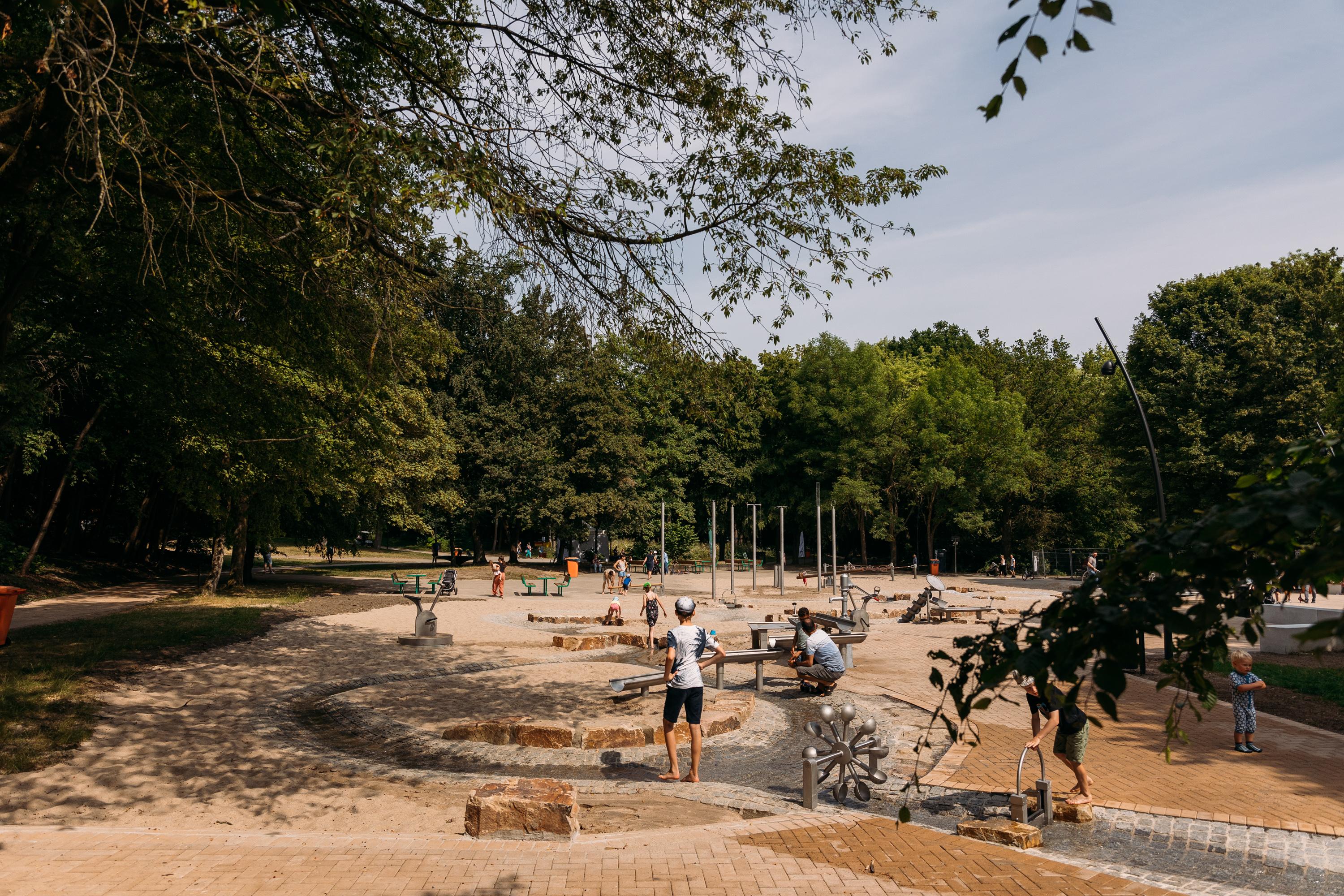 An einem von Bäumen eingefassten Platz spielen Kinder auf einem Wassespielplatz.