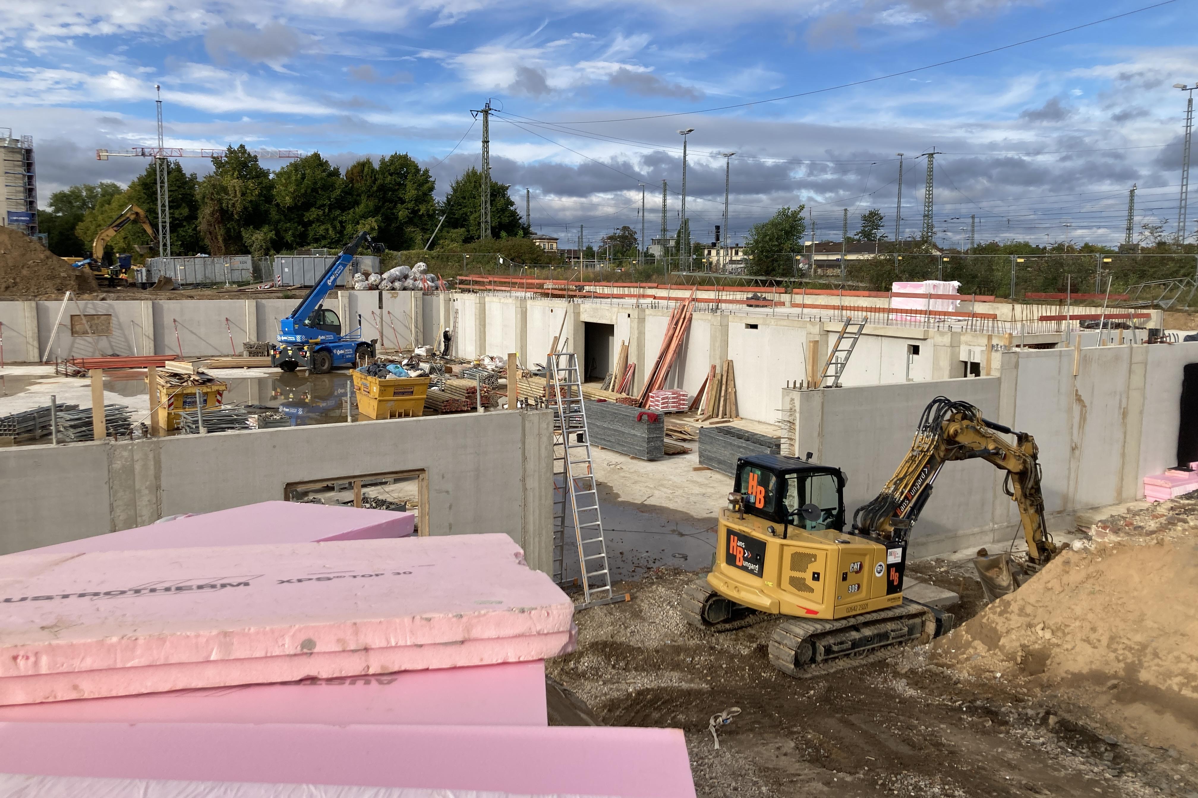 Baustelle mit Baggern, Baukränen und unfertigen Betonwänden; im Vordergrund liegt pinke Dämmung, während Arbeiter und Maschinen im Hintergrund am Rohbau arbeiten.