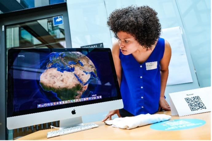 Vor einem Computerbildschirm auf einem Schreibtisch lehnt eine weibliche Person, um den Inhalt des Bildschirmes zu sehen. 