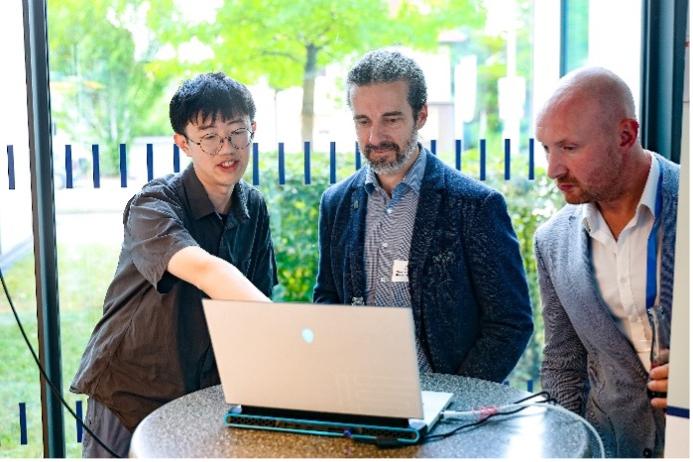 Drei Personen stehen um einen Stehtisch mit einem Laptop. Eine Person zeigt auf den Bildschirm des Laptops. 