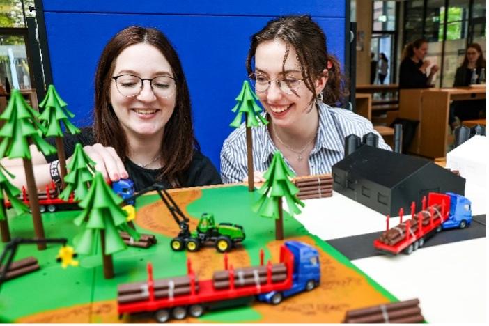 Zwei Personen schauen auf einen Tisch mit exemplarisch gefertigten Fahrzeugen, Bäumen und Hallen.