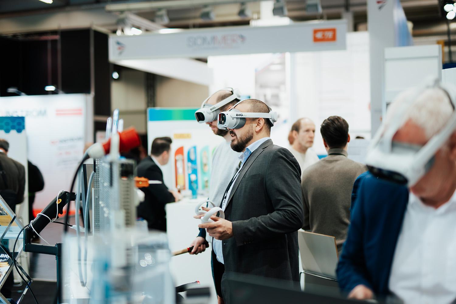 Zwei Personen mit VR-Brillen an einem Stand auf einer Messe. 