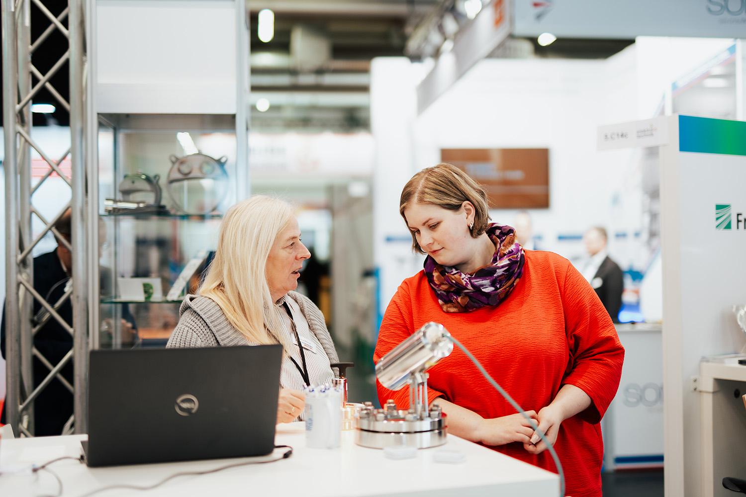 Zwei Personen stehen an einem Tisch und schauen sich einen Roboterarm auf einer Messean. 