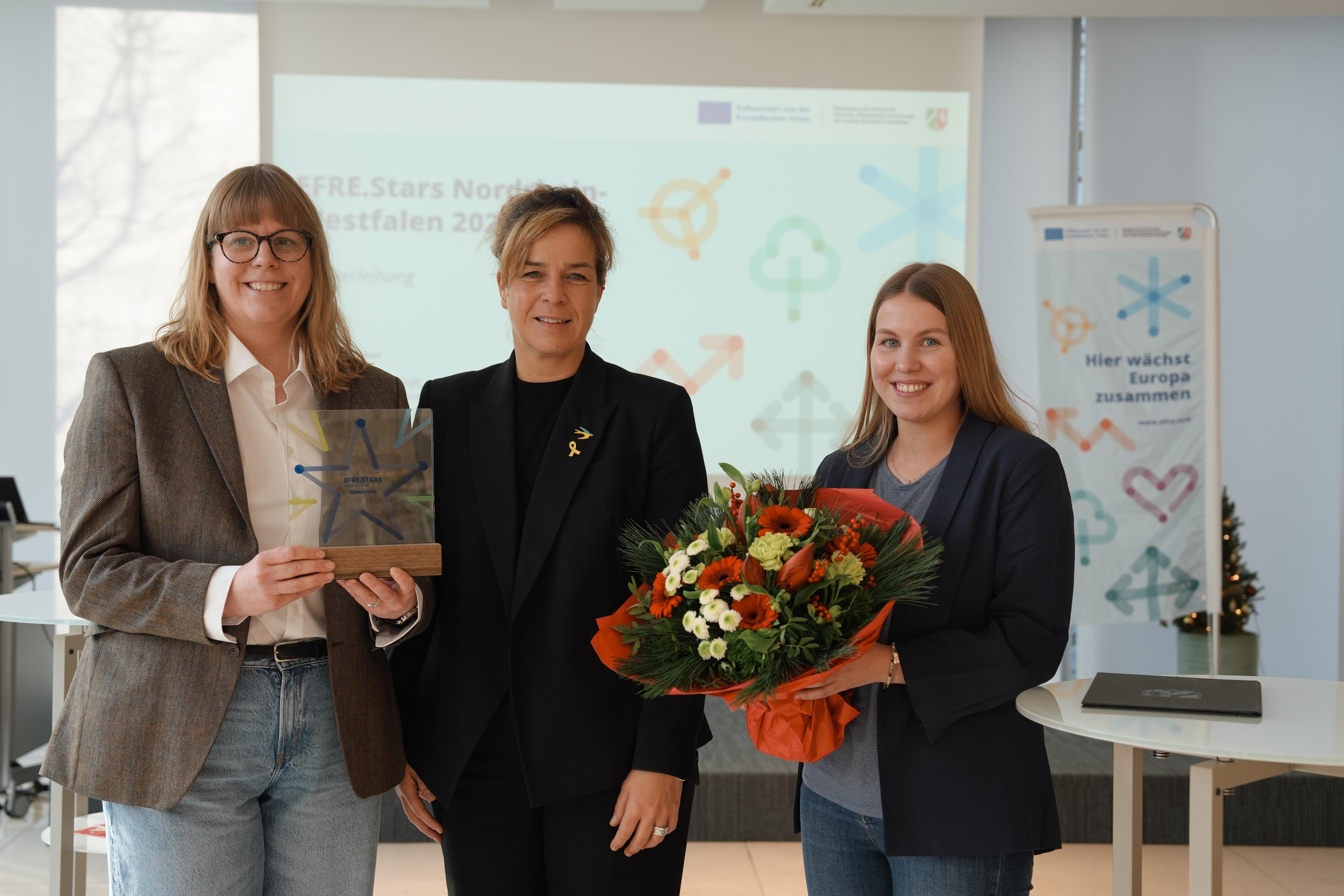 Drei Personen schauen lächelnd in die Kamera. Eine Person hält einen Preis in der Hand, eine andere einen Blumenstrauß.