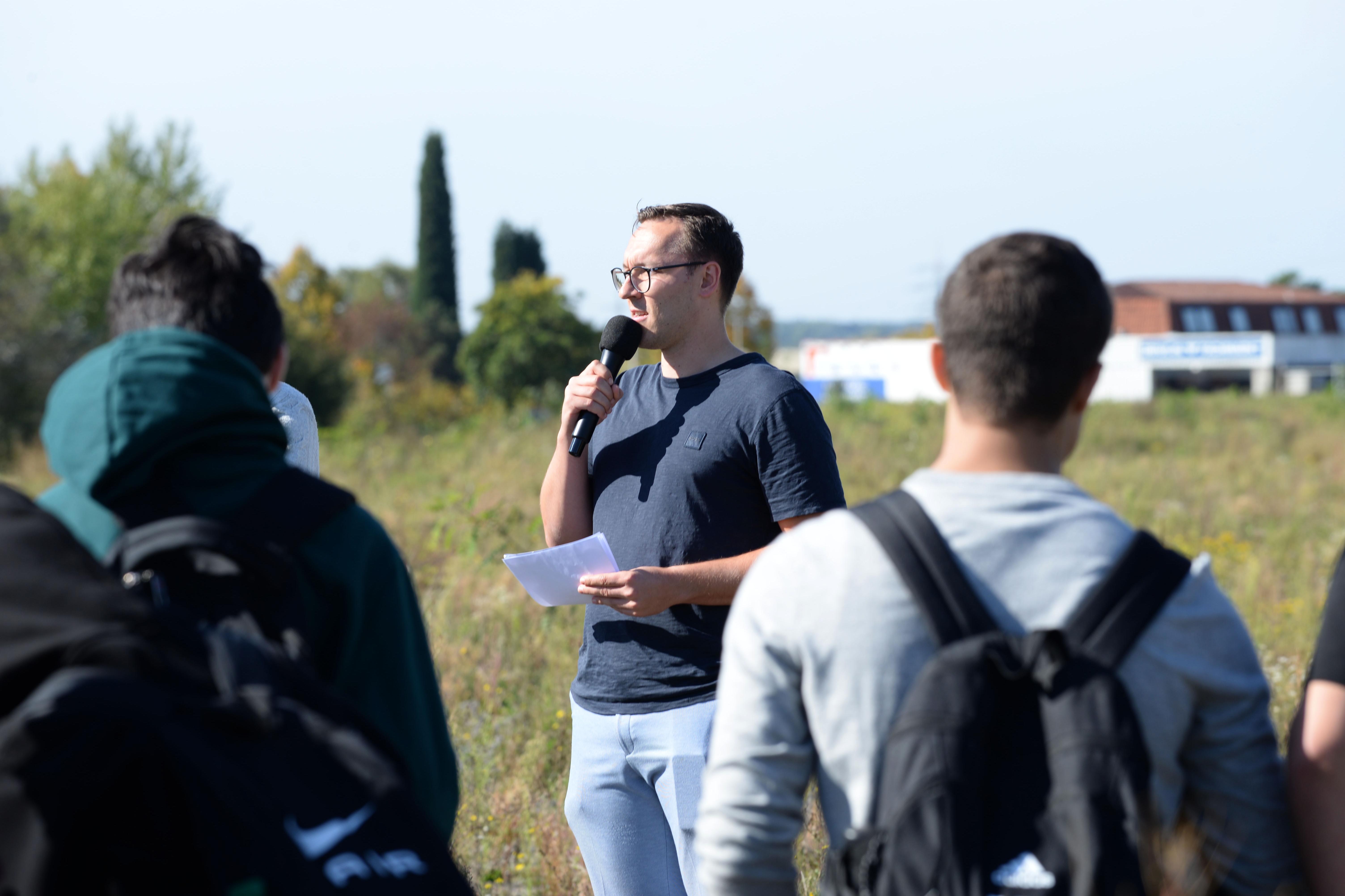 Ein Mann spricht in ein Mikrofon und erzählt eine Gruppe von Menschen etwas.