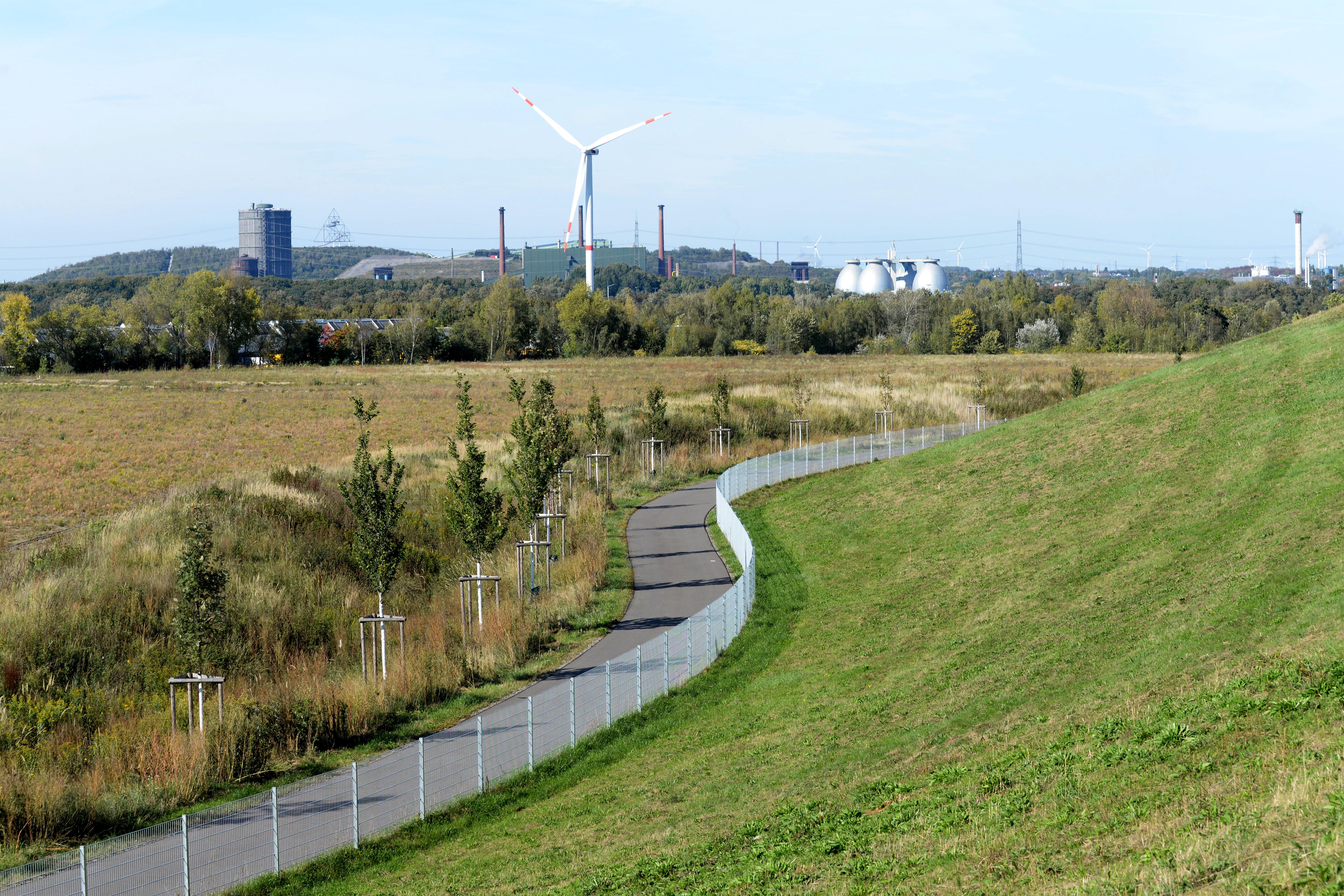 Es ist ein Weg inmitten einer Landschaft zu sehen. Im Hintergrund ist eine Industrielandschaft zu sehen.