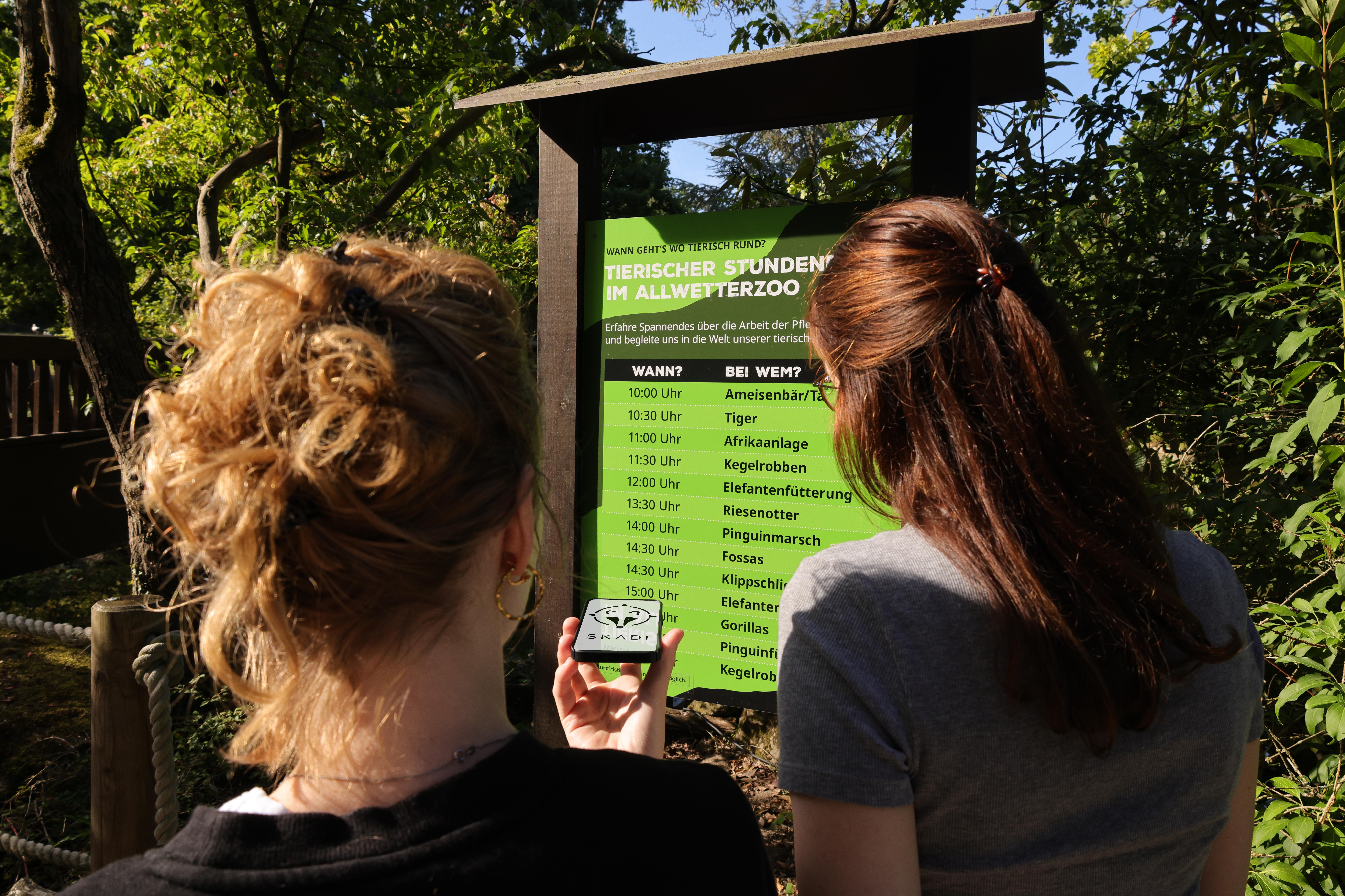 Zwei Besucherinnen im Allwetterzoo Münster vor einer Tafel mit Zeitangaben