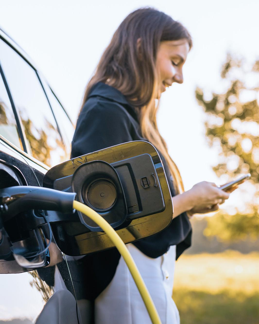 Eine Frau steht ans Auto gelehnt, die Ladeklappe ist offen und ein Kabel steckt am Auto