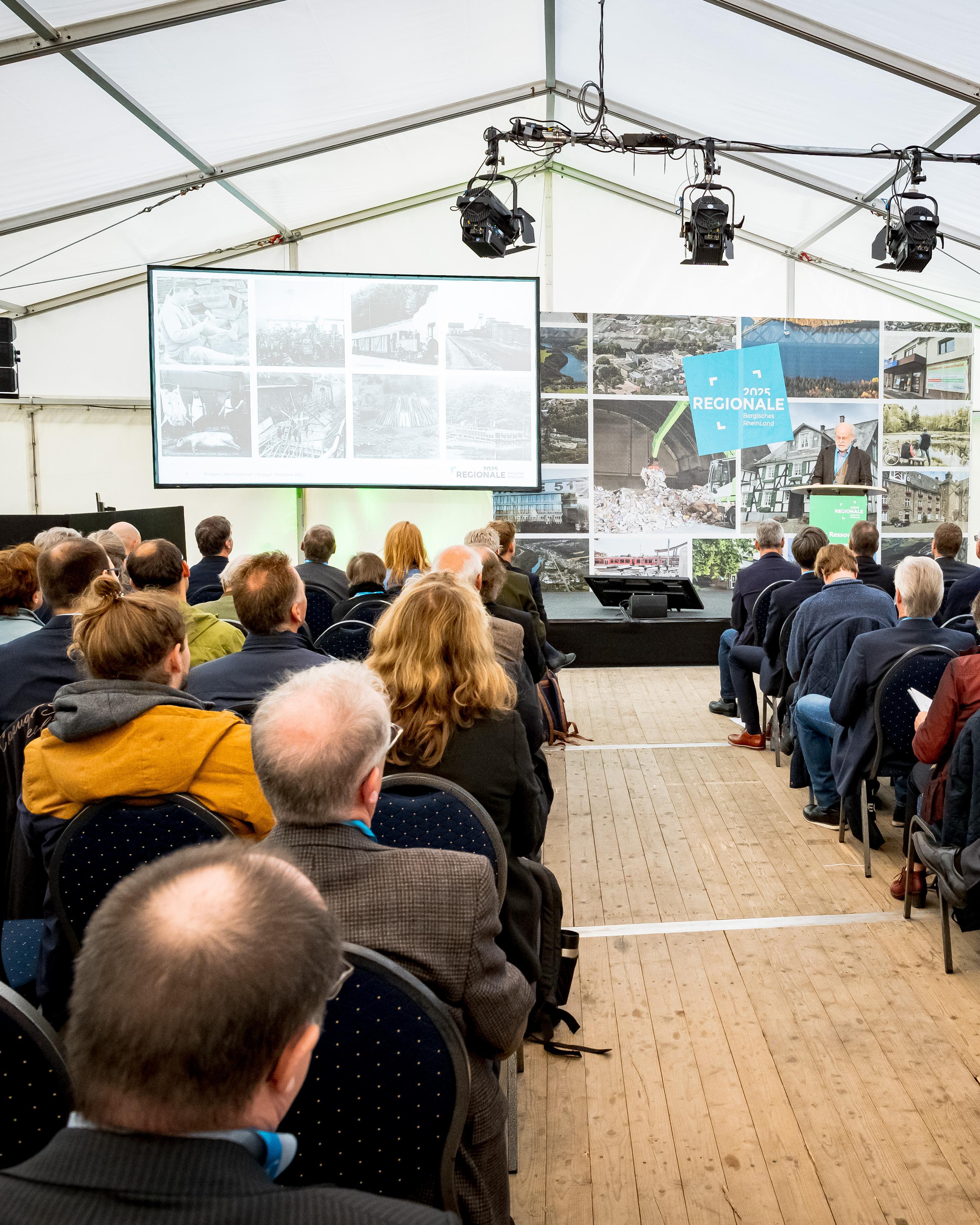 Es ist ein Konferenzraum mit Stuhlreihen abgebildet, auf denen viele Menschen sitzen.