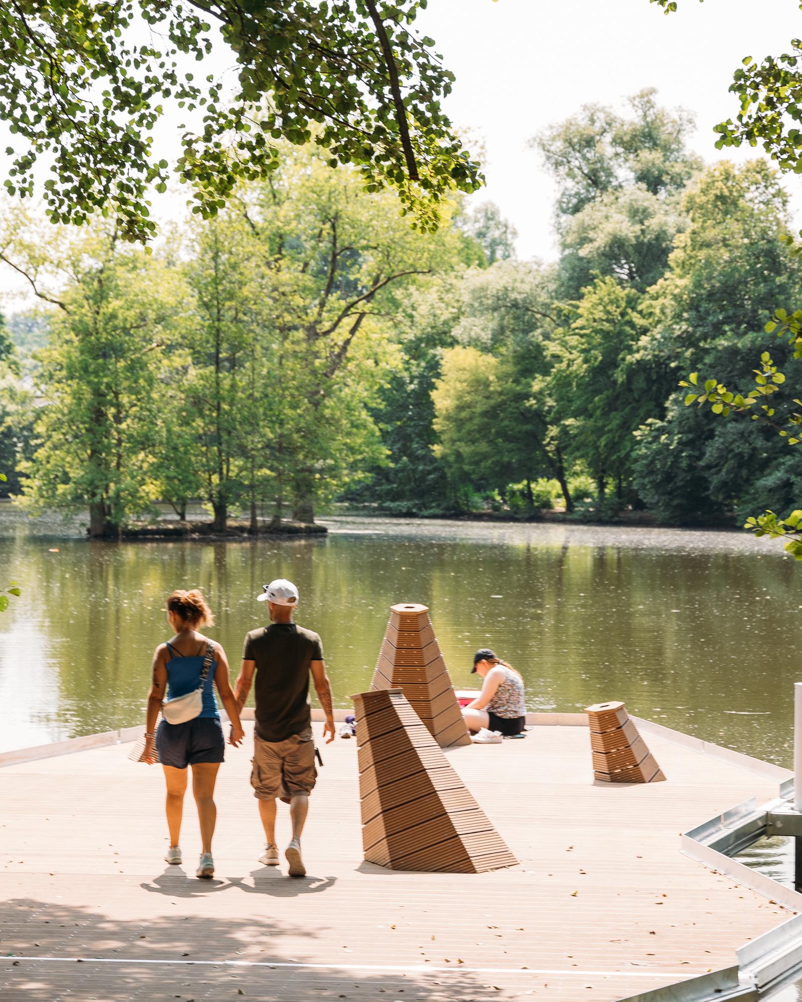 Auf einer kleinen, schwimmenden Plattform in einem See, um dessen Ufer herum Bäume wachsen, sitzt eine Person direkt am Wasser. Zwei weitere Personen gehen Hand in Hand über die Plattform und schauen auf den See.