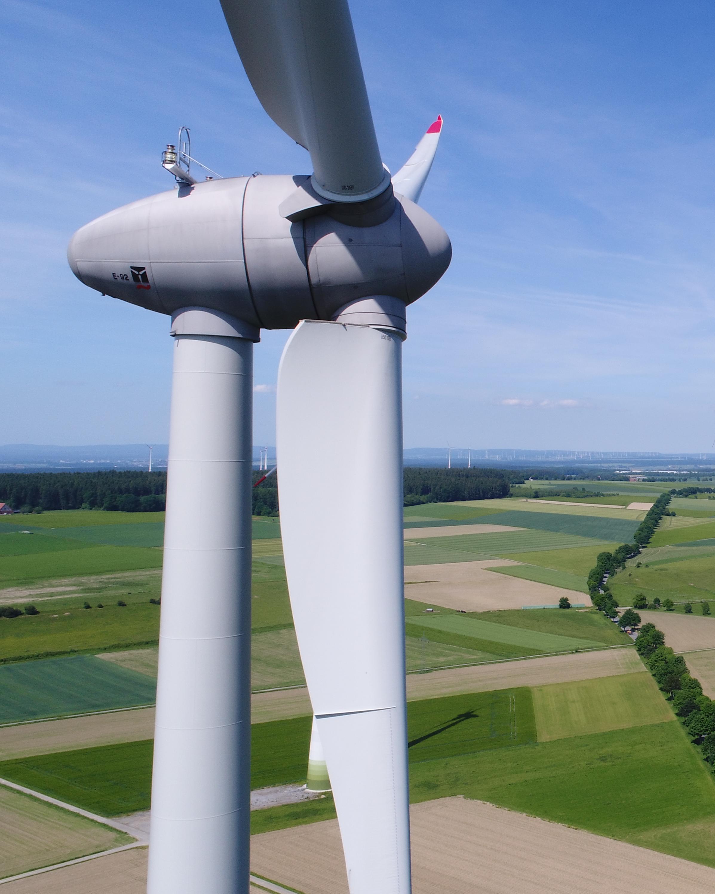 Ein Windrad steht in einer freien Fläche grüner Felder. Der Himmel ist blau, am Horizont sind weitere Windräder zu erkennen.