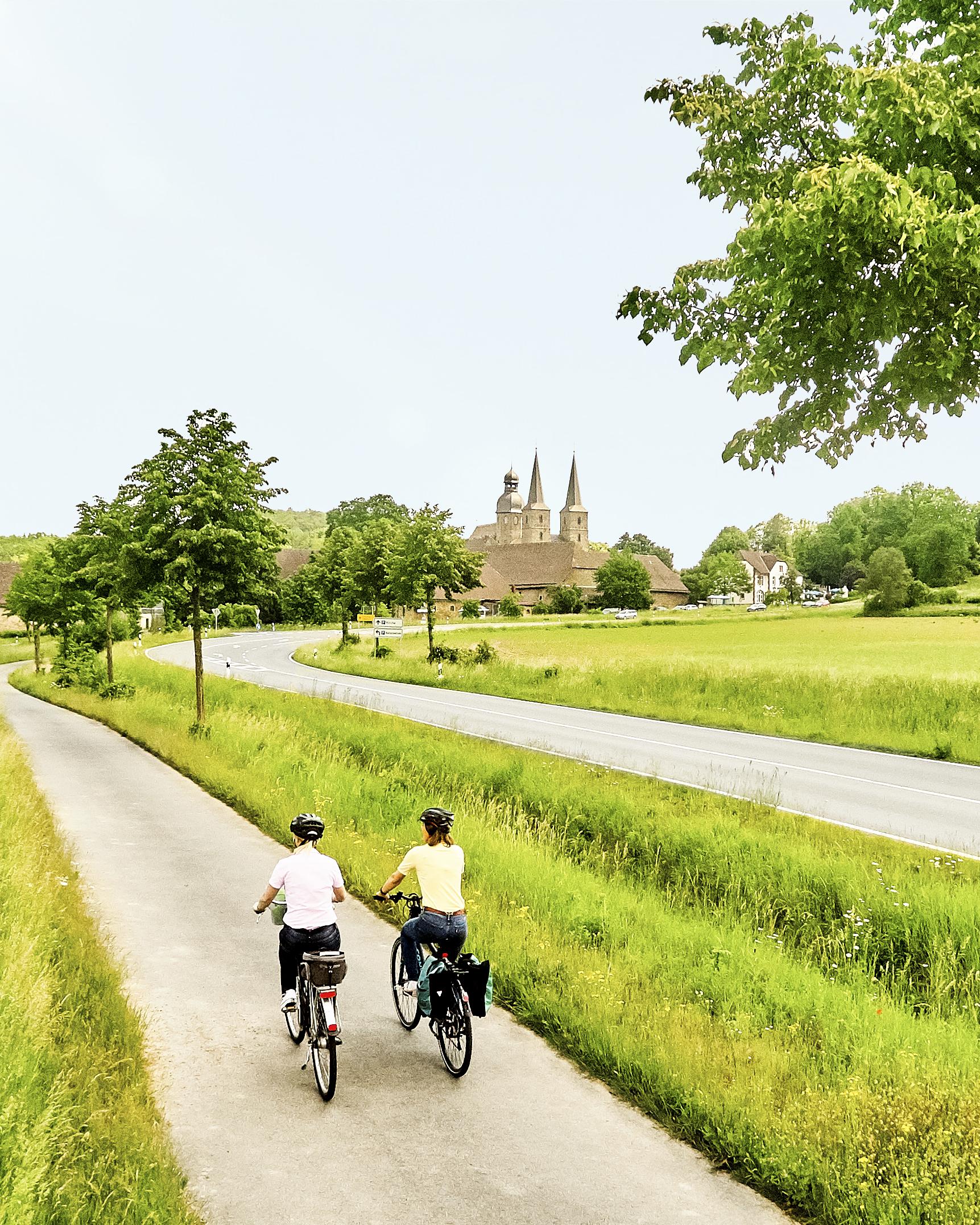 Ein Fahrradweg führt an einer Landstraße entlang über grüne Felder. Auf dem Weg radeln zwei Menschen, am Horizont ein Dorf. 