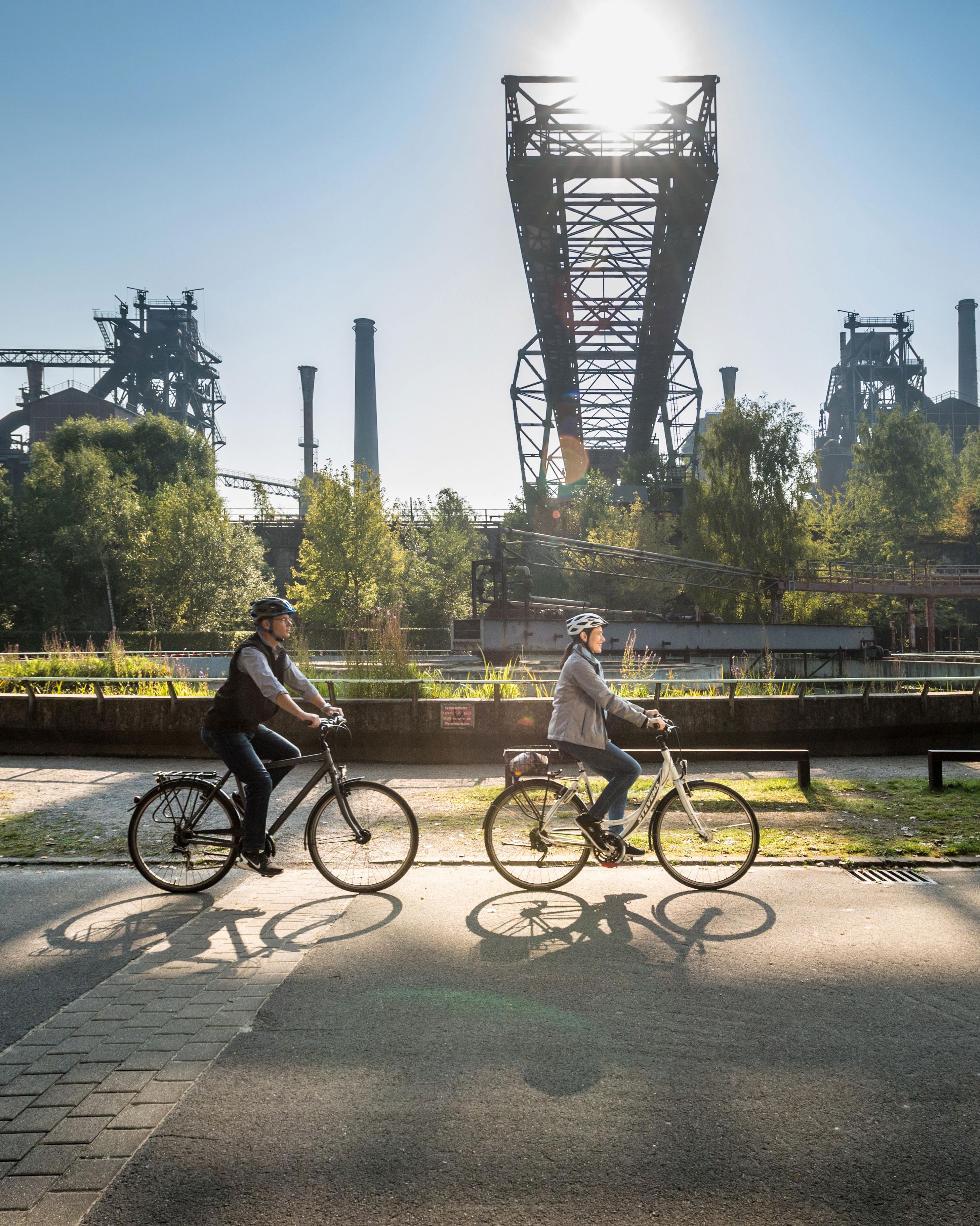 Zwei Personen radeln auf einem Fahrradweg, der an einer alten Industrieanlage vorbeiführt.