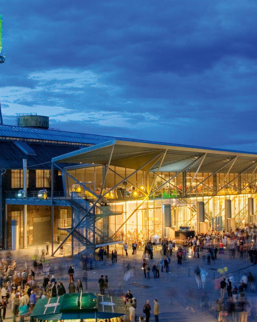 Die Jahrhunderhalle bei Nacht. Das Gebäude ist hell beläuchtet, davor halten sich viele Menschen auf.