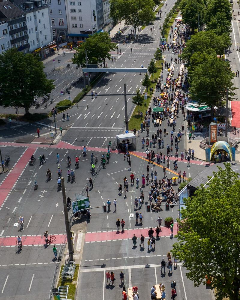 Eine belebte Straßenkreuzung aus der Vogelperspektive. Auf der Straße halten sich viele Menschen auf, während keine Autos unterwegs sind.