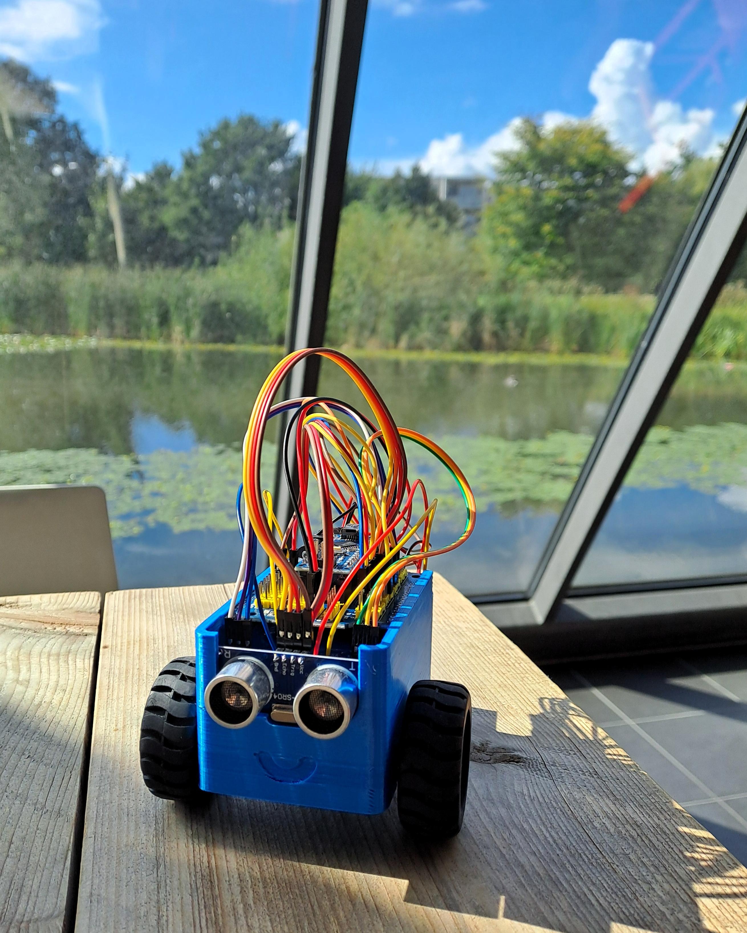 Es ist ein 3D-gedruckter kleiner Roboter aus dem viele bunte Kabel hervorkommen vor einer Glasfront mit Blick auf einen See zu sehen.