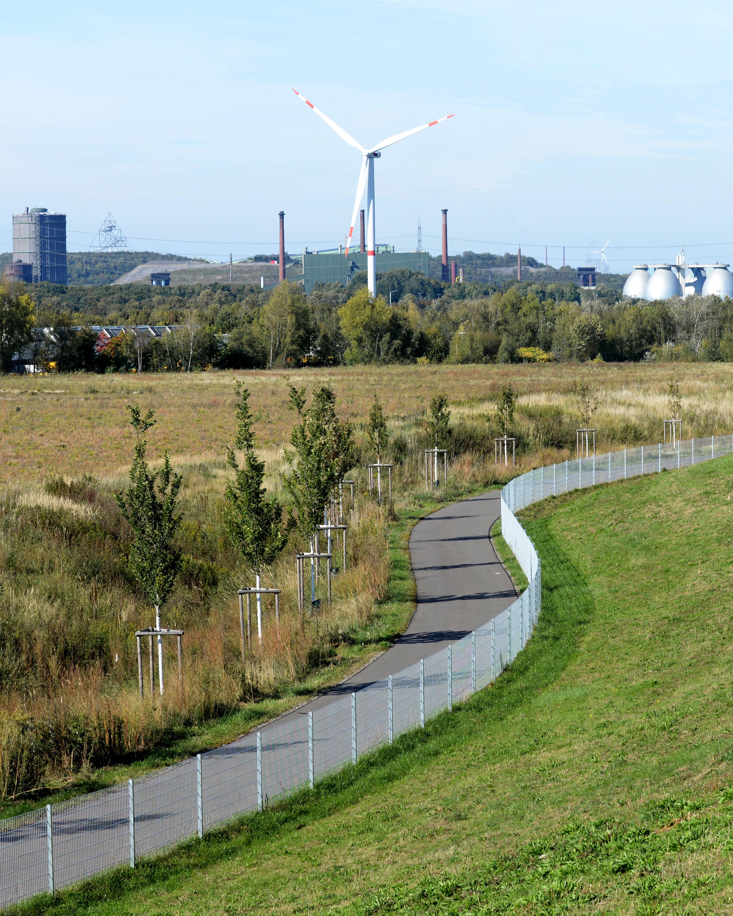 Es ist ein Weg inmitten einer Landschaft zu sehen. Im Hintergrund ist eine Industrielandschaft zu sehen.