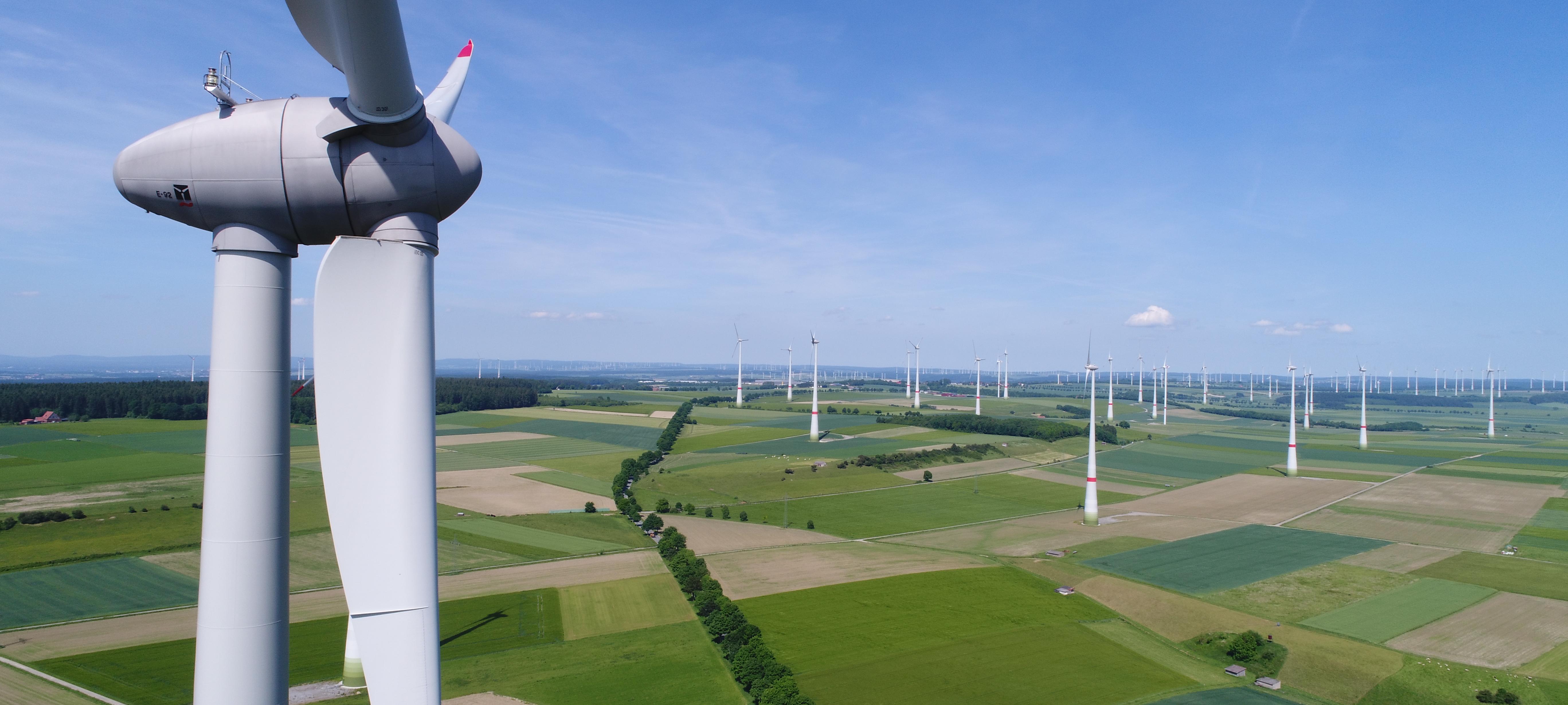 Ein Windrad steht in einer freien Fläche grüner Felder. Der Himmel ist blau, am Horizont sind weitere Windräder zu erkennen.