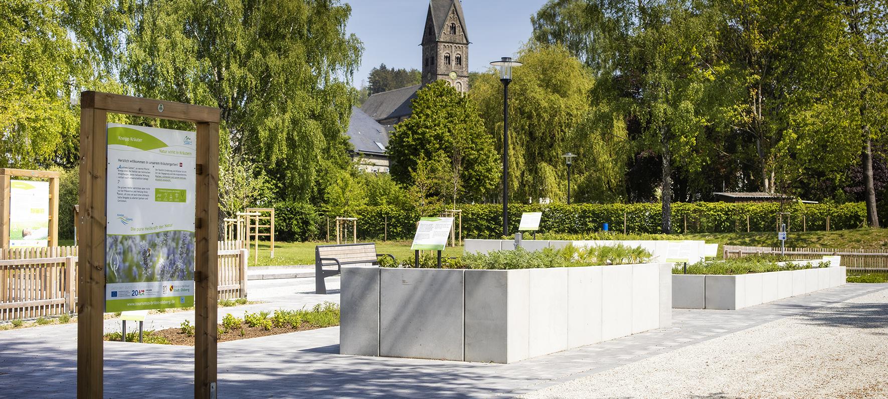 Ein begrünter Dorfplatz, von Bäumen umringt und mit Beeten in verschiedenen Höhen. Im Hintergrund ragt ein Kirchturm hervor. Vor dem Platz steht ein Infoschild.