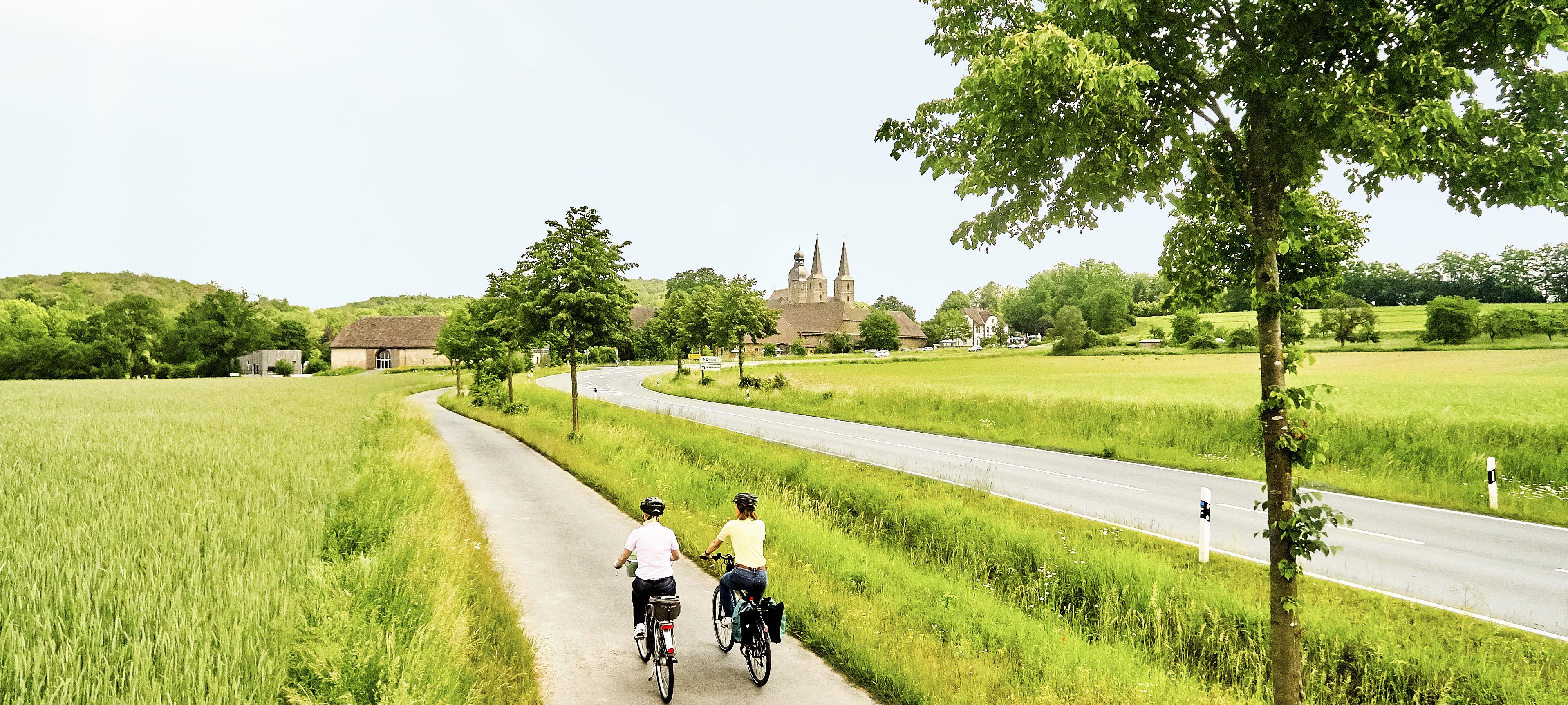 Ein Fahrradweg führt an einer Landstraße entlang über grüne Felder. Auf dem Weg radeln zwei Menschen, am Horizont ein Dorf. 