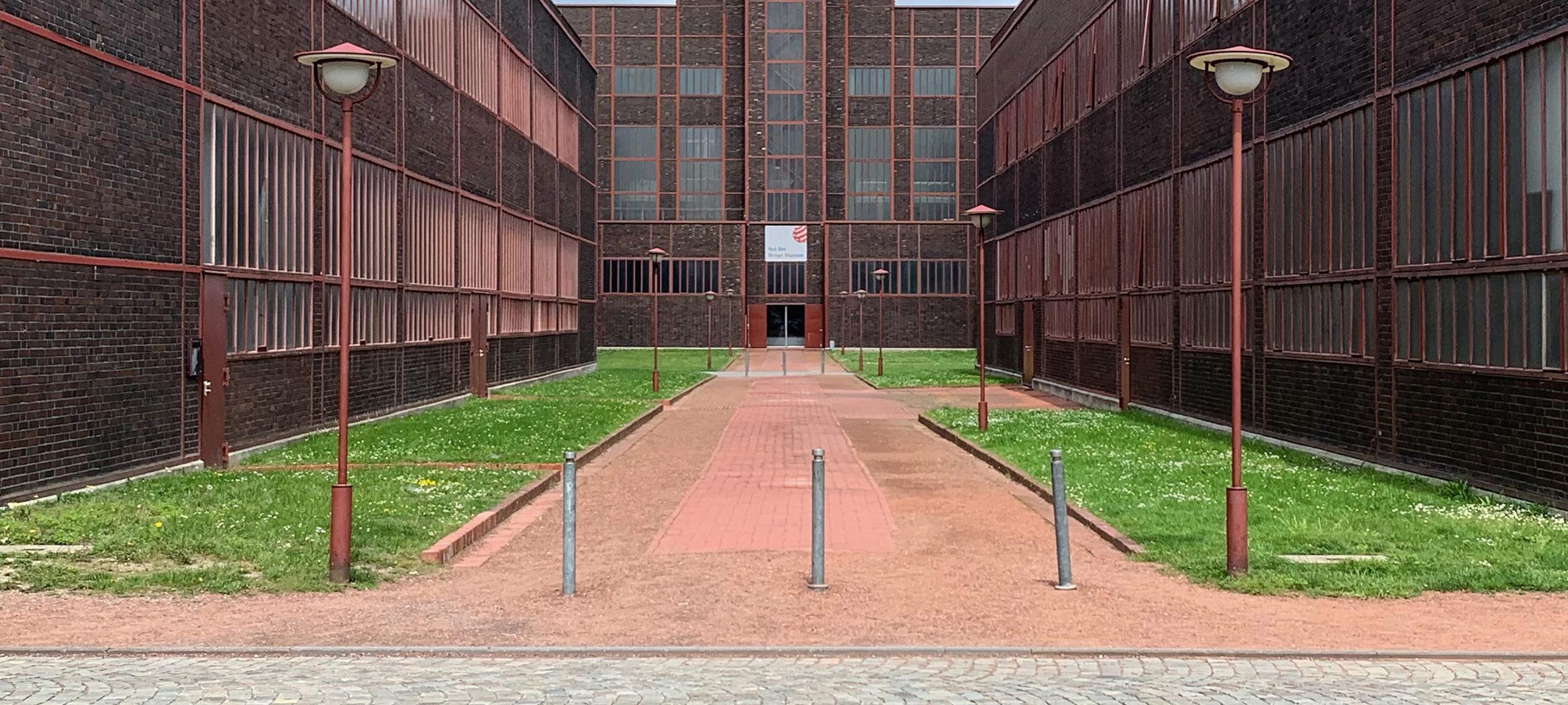 Ein Klinkerbau von außen mit roten Details und Straßenlaternen. Zwischen den einzelnen Gebäudeteilen verlaufen begrünte Fußwege.