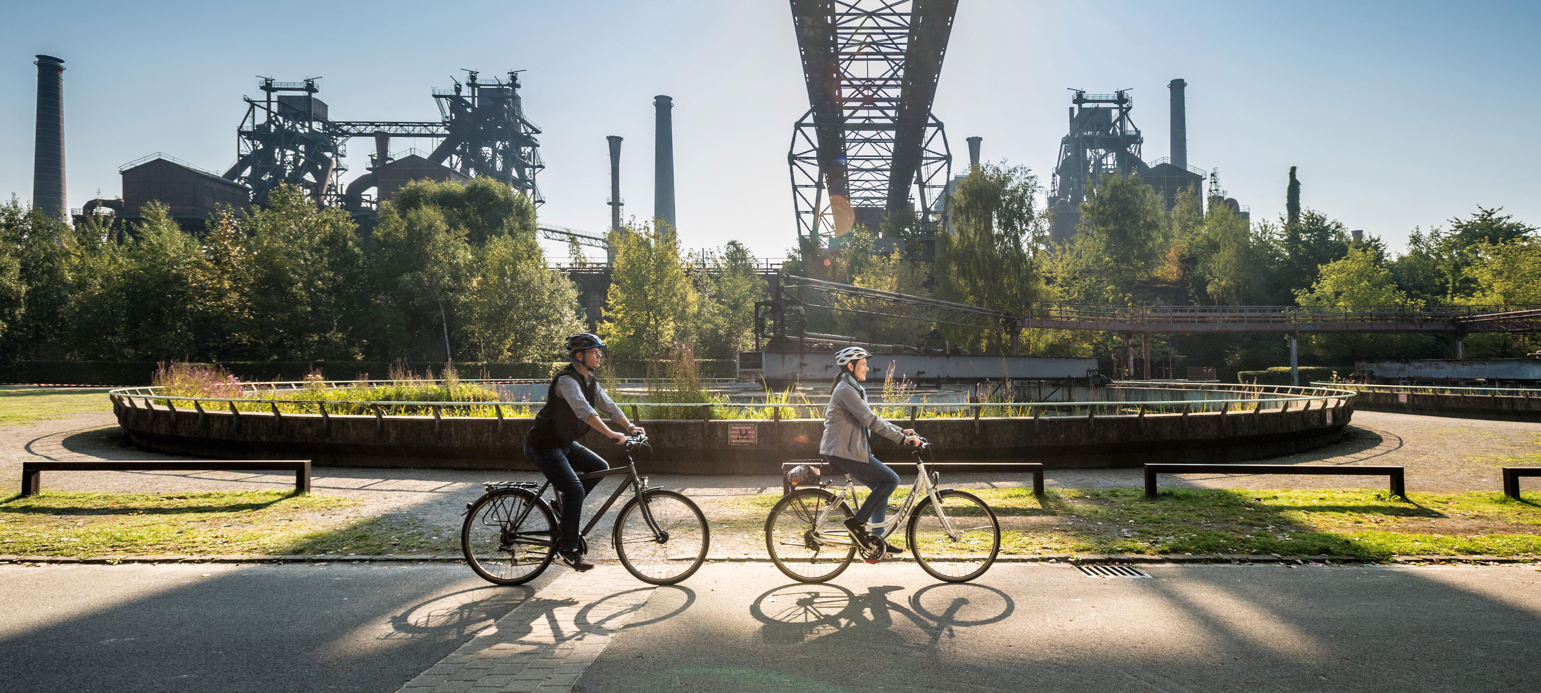 Zwei Personen radeln auf einem Fahrradweg, der an einer alten Industrieanlage vorbeiführt.