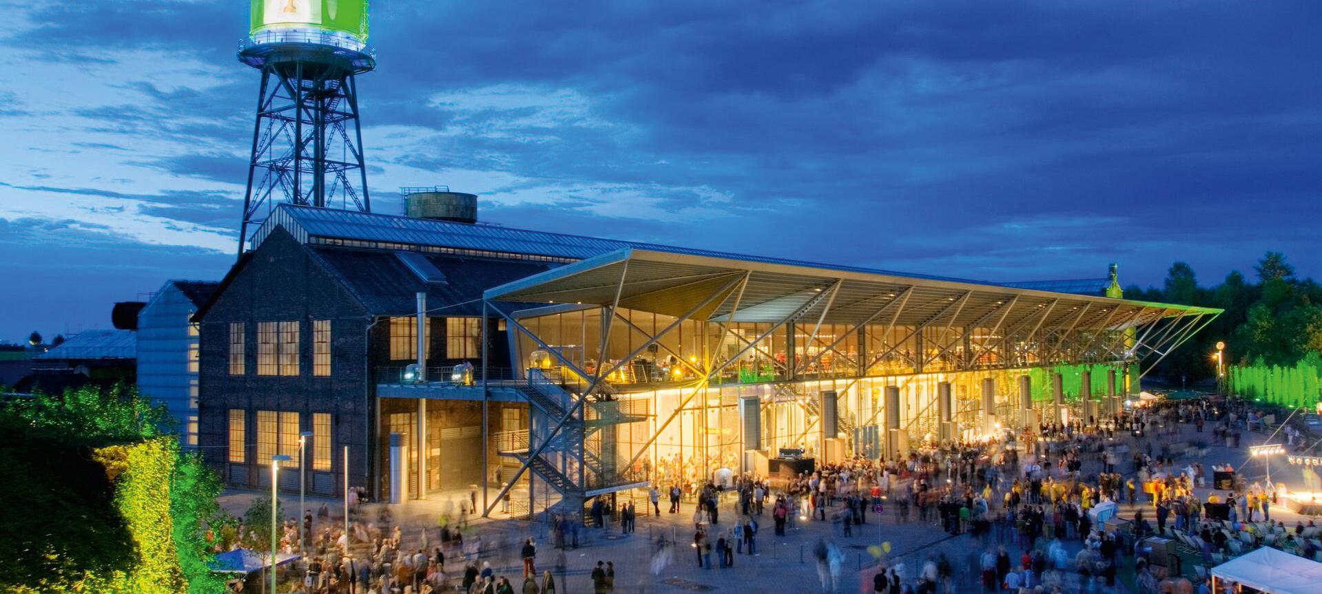 Die Jahrhunderhalle bei Nacht. Das Gebäude ist hell beläuchtet, davor halten sich viele Menschen auf.