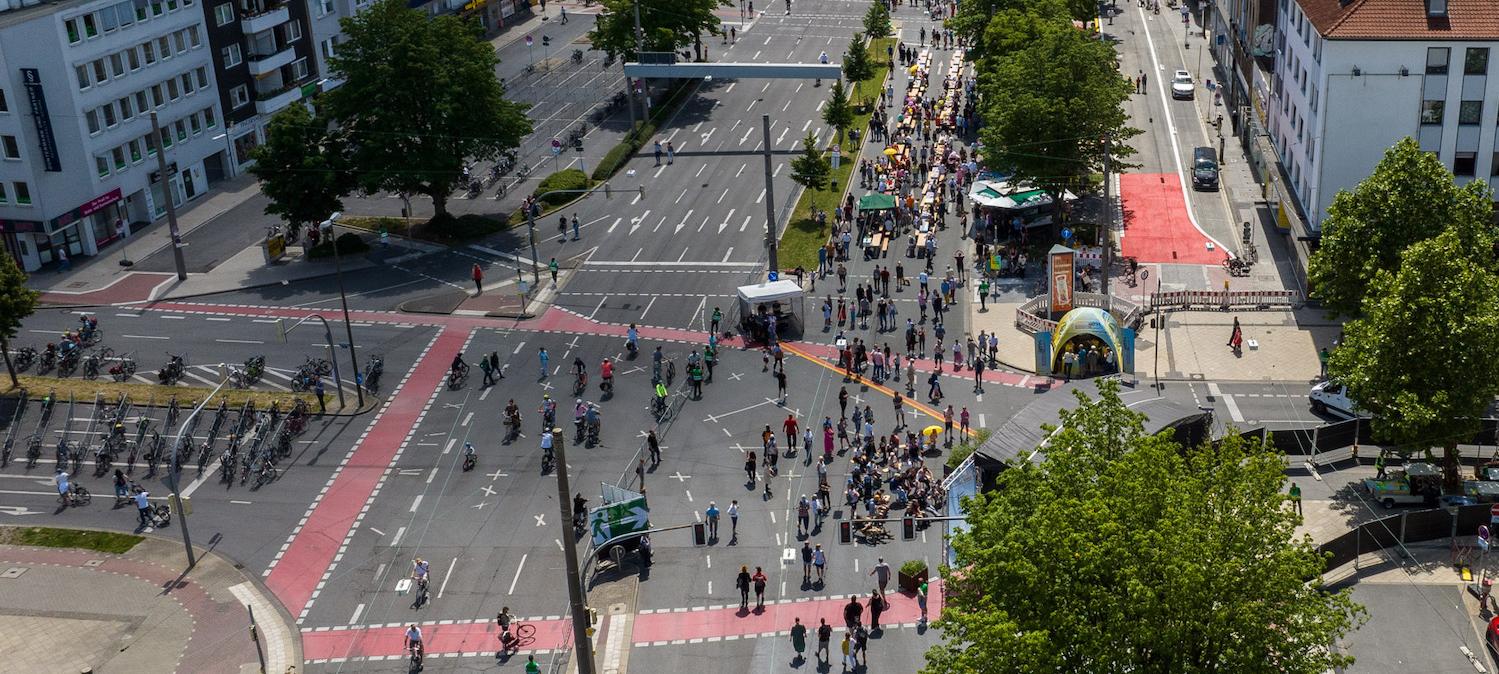 Eine belebte Straßenkreuzung aus der Vogelperspektive. Auf der Straße halten sich viele Menschen auf, während keine Autos unterwegs sind.