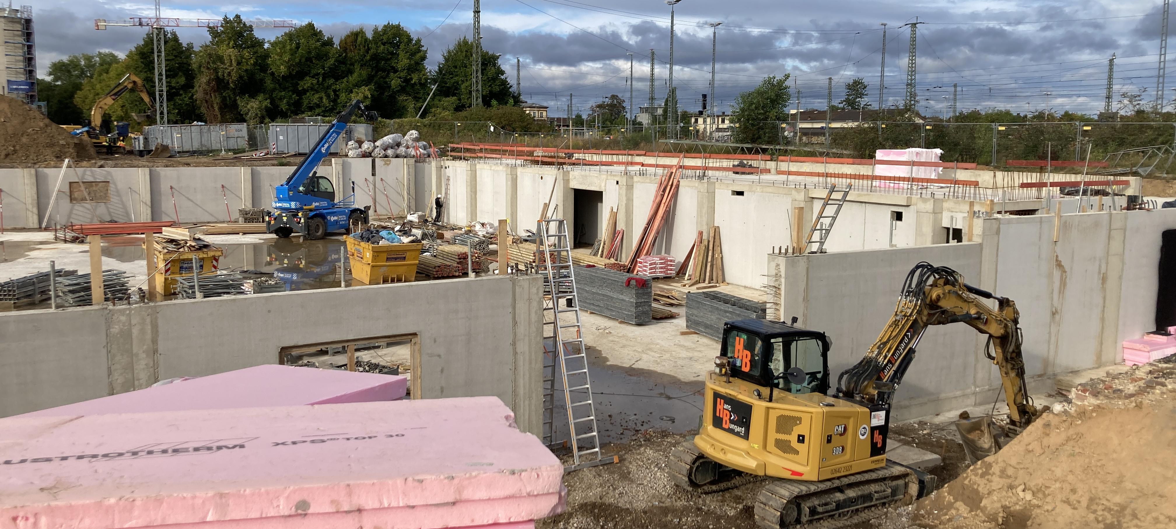 Baustelle mit Baggern, Baukränen und unfertigen Betonwänden; im Vordergrund liegt pinke Dämmung, während Arbeiter und Maschinen im Hintergrund am Rohbau arbeiten.