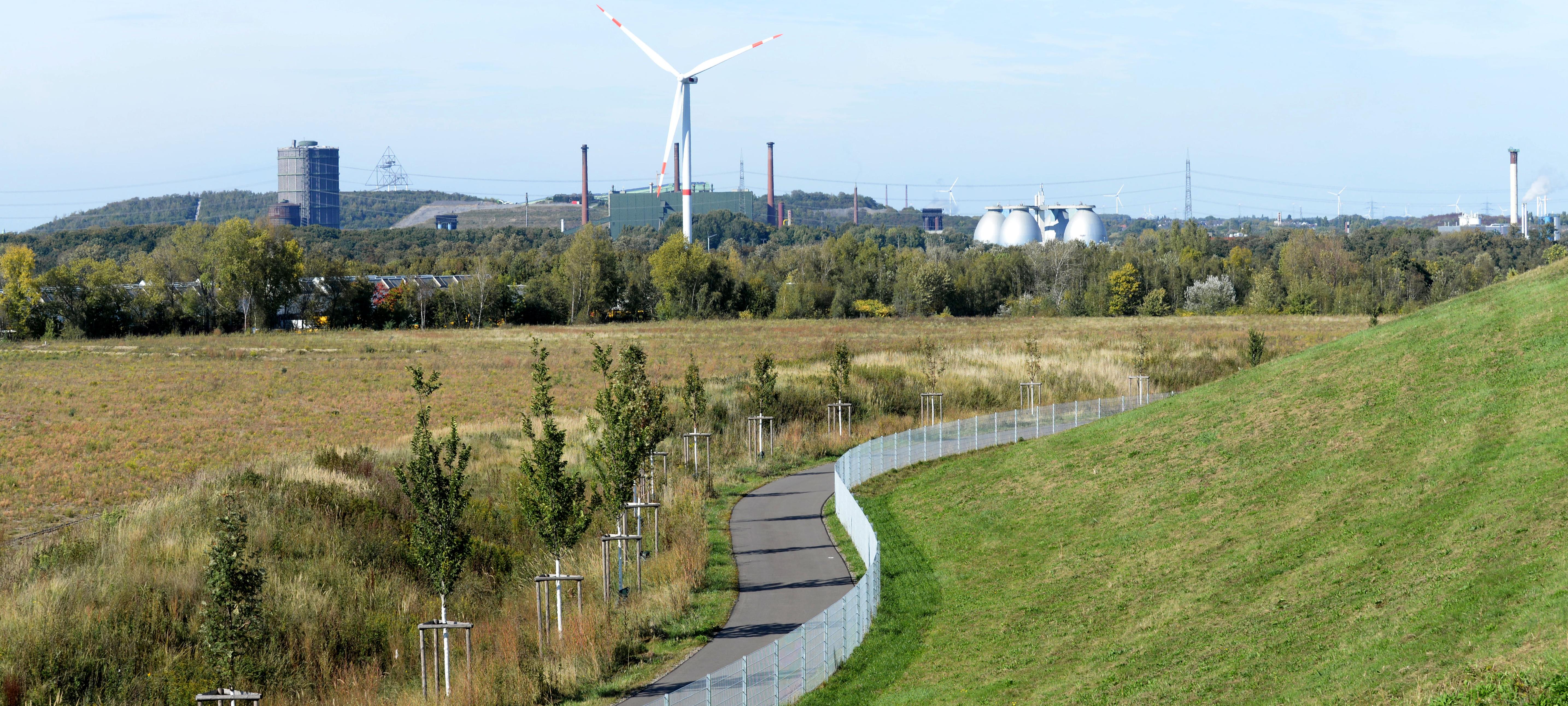 Es ist ein Weg inmitten einer Landschaft zu sehen. Im Hintergrund ist eine Industrielandschaft zu sehen.
